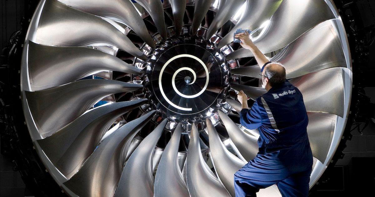 A technician in a blue uniform inspects or polishes the large, shiny fan blades of a Rolls-Royce aircraft engine. The fan has a prominent spiral design at its center.