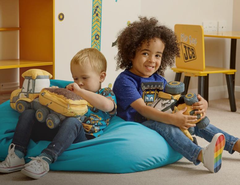 Two young children sit on a blue bean bag in a playroom, smiling and holding soft toy construction vehicles. A yellow desk and wall decorations are visible in the background.