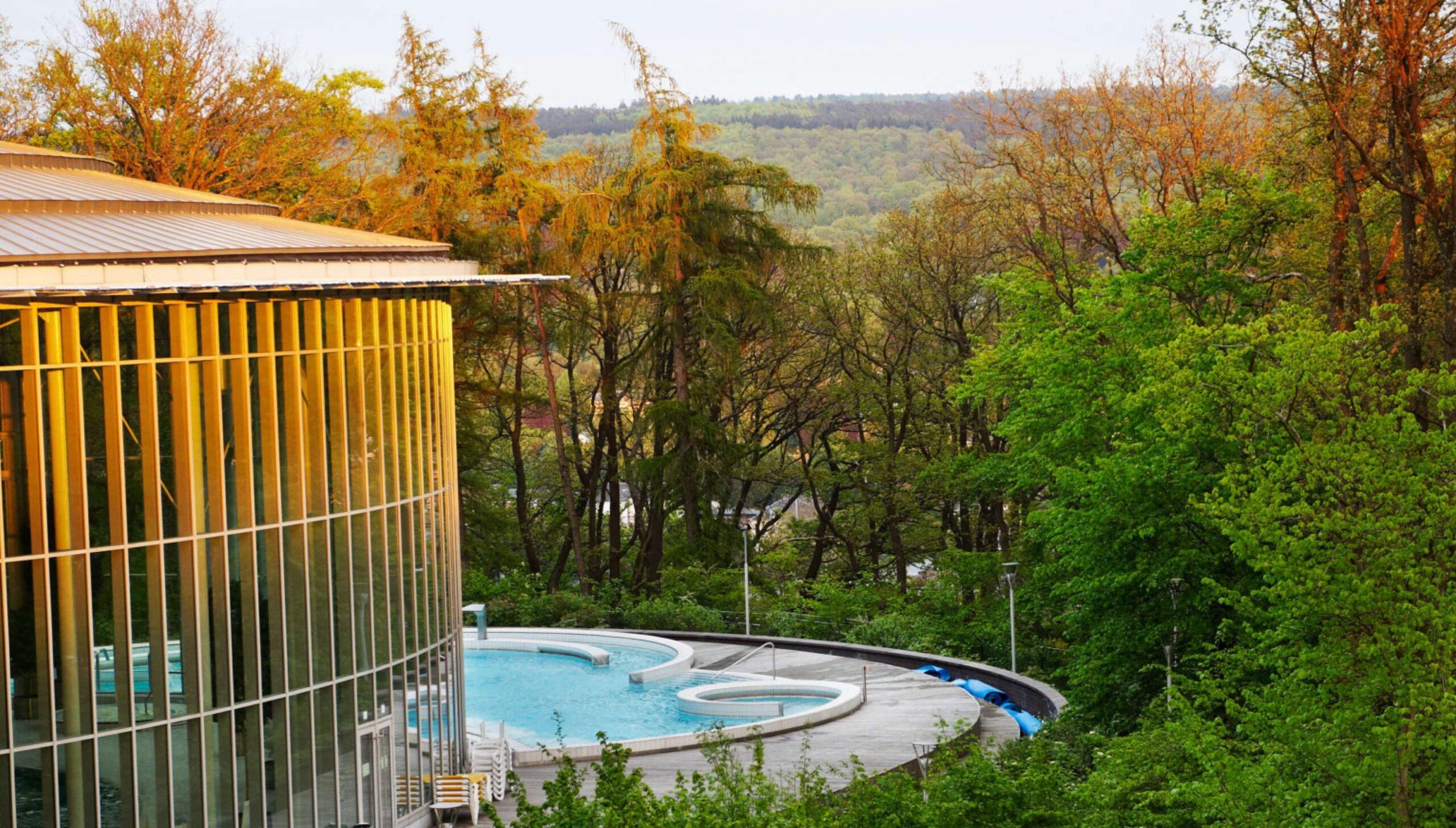 A modern, glass-walled building with an outdoor circular pool is surrounded by lush green trees and forested hills under a clear sky at sunset.