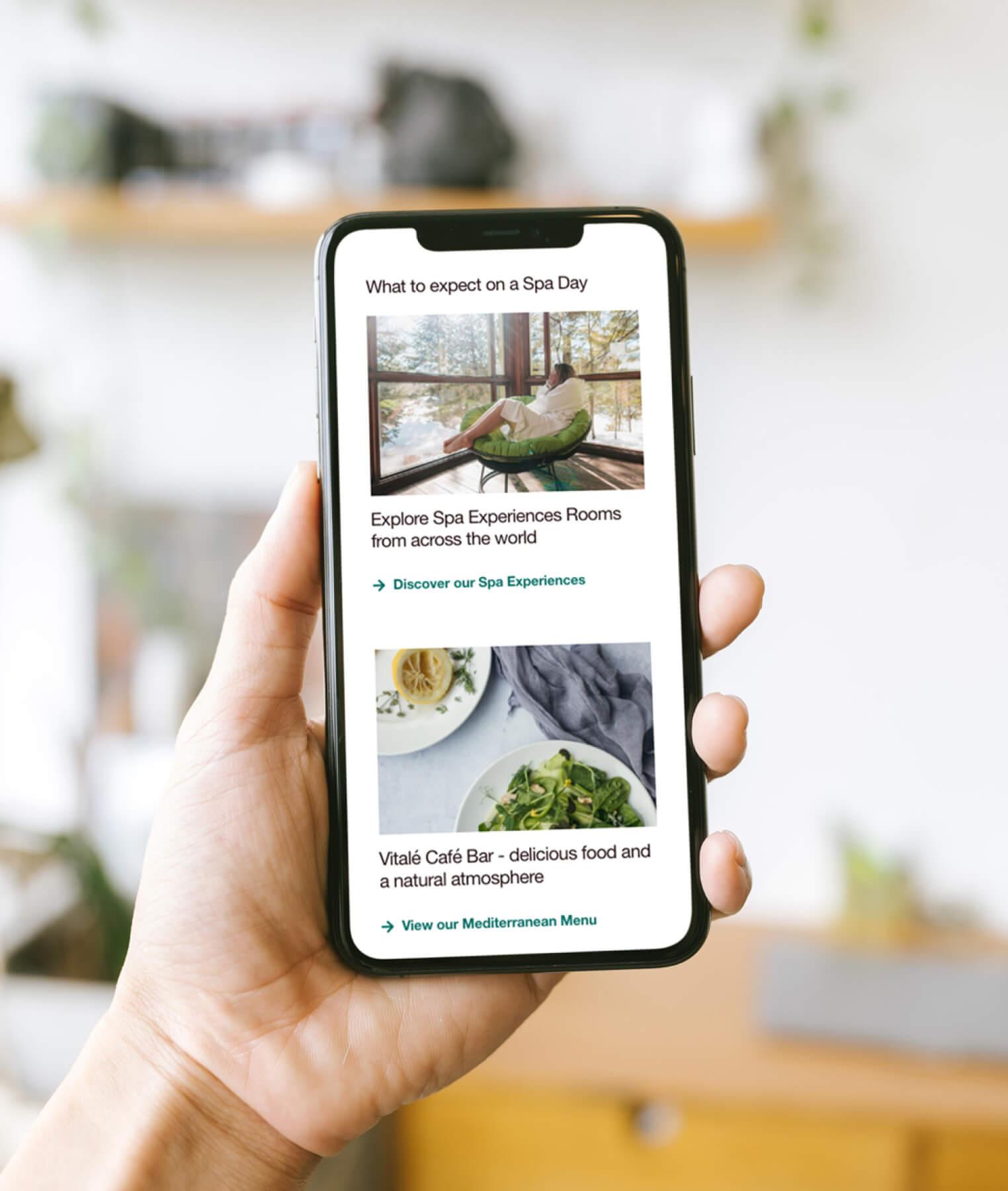 A hand holds a smartphone displaying a website about spa days, featuring images of a relaxing lounge chair by a window and a healthy meal, with text describing spa experiences and a Mediterranean café bar.