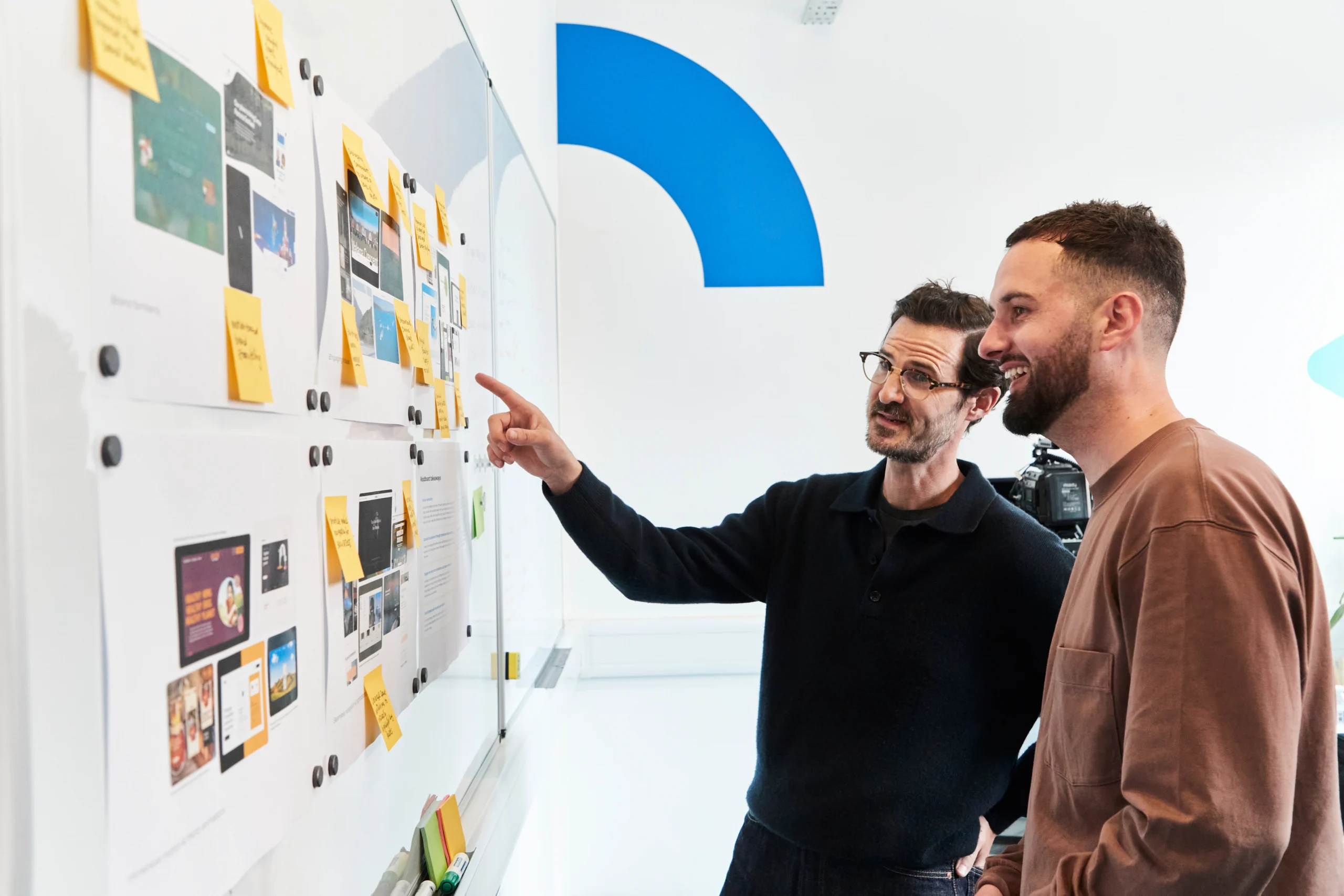 Two men standing at a whiteboard covered with papers and sticky notes, discussing and pointing at the materials, appearing engaged in a collaborative brainstorming or planning session in a modern office.