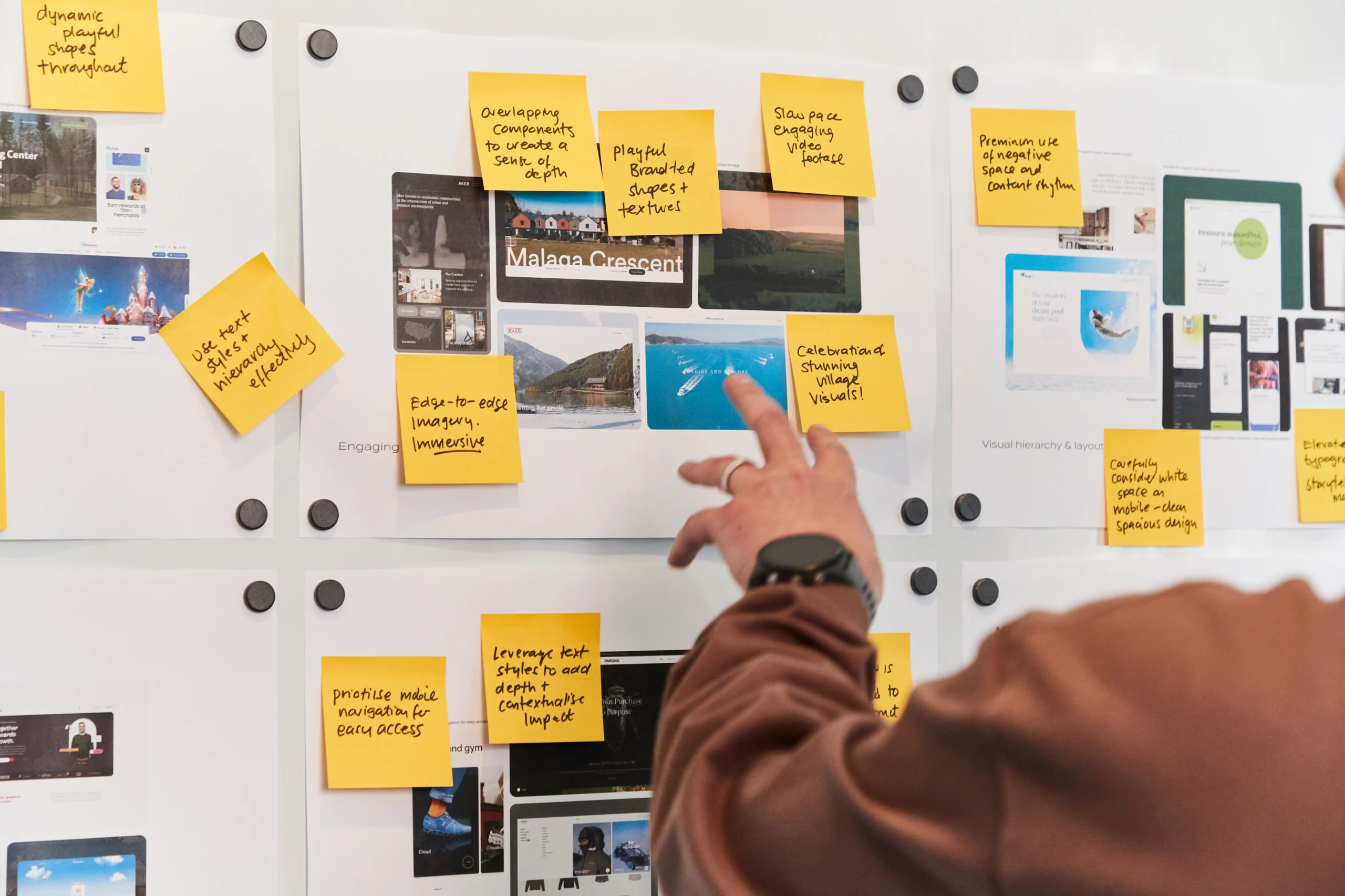 Person pointing at a whiteboard covered with website screenshots and yellow sticky notes containing handwritten ideas, tasks, and observations for a project or design brainstorming session.