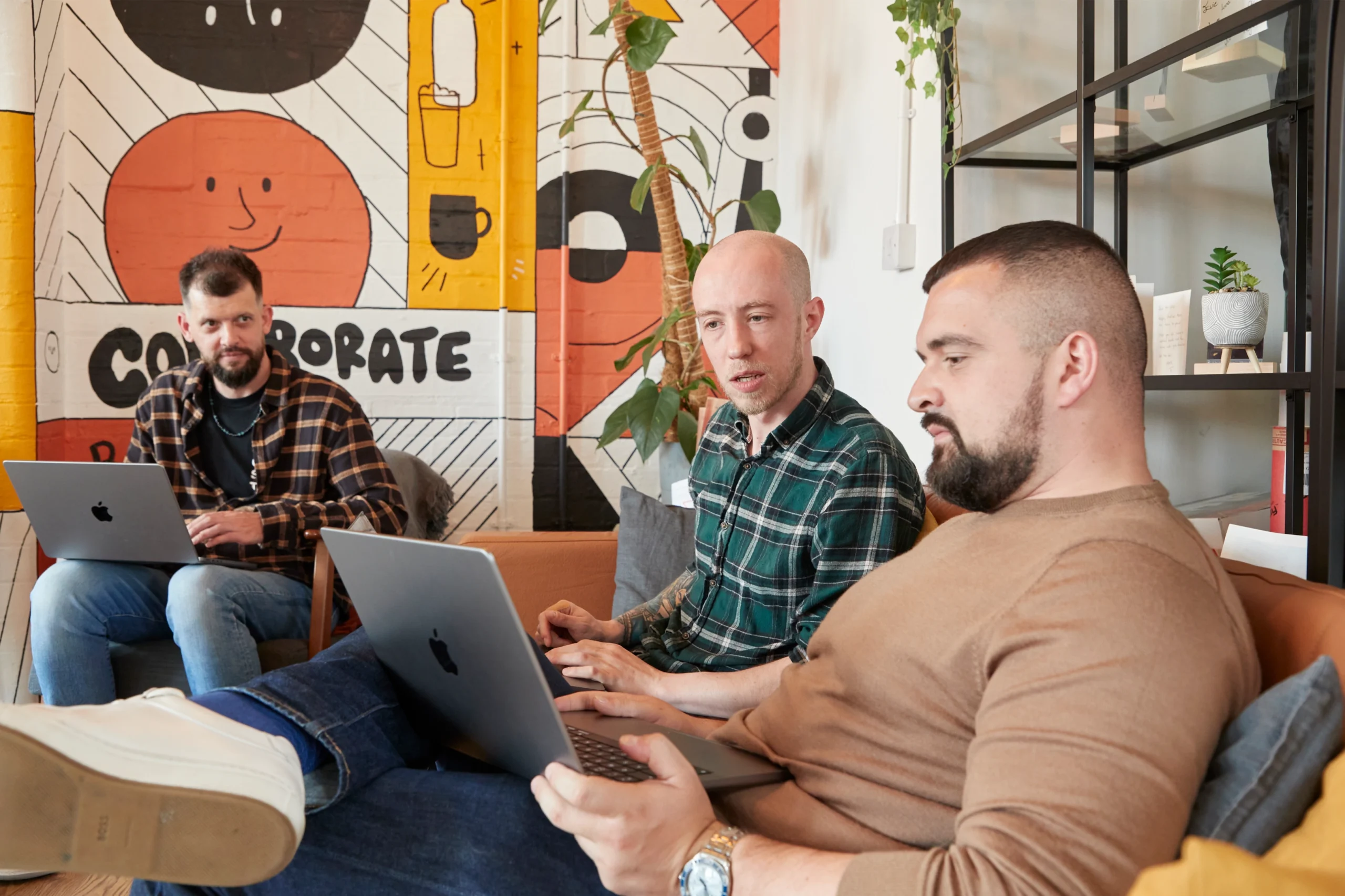 Three men sit on a sofa with laptops, engaged in discussion. A colourful mural with abstract faces and CORPORATE text is on the wall behind them. Shelves with plants are visible to the right.