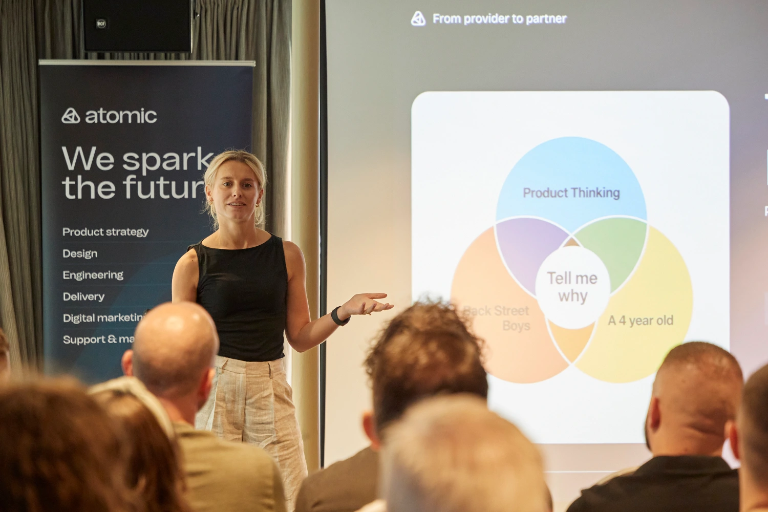 A woman gives a presentation to an audience, standing next to a screen displaying a colourful Venn diagram and a banner reading atomic: We spark the future with a list of services.