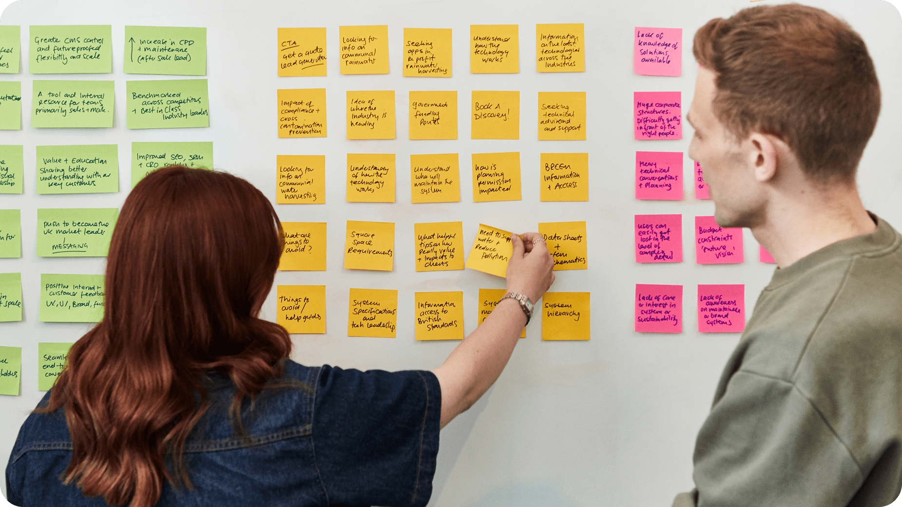 Two people stand in front of a whiteboard filled with colorful sticky notes. One person is placing a note on the board while the other observes, suggesting a collaborative brainstorming session.