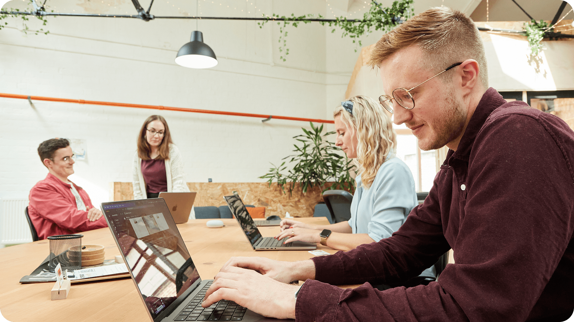 Four people work together in a modern office. Two sit at a table typing on laptops, while another sits nearby and one stands, looking at a screen. The space is bright with plants and natural light.