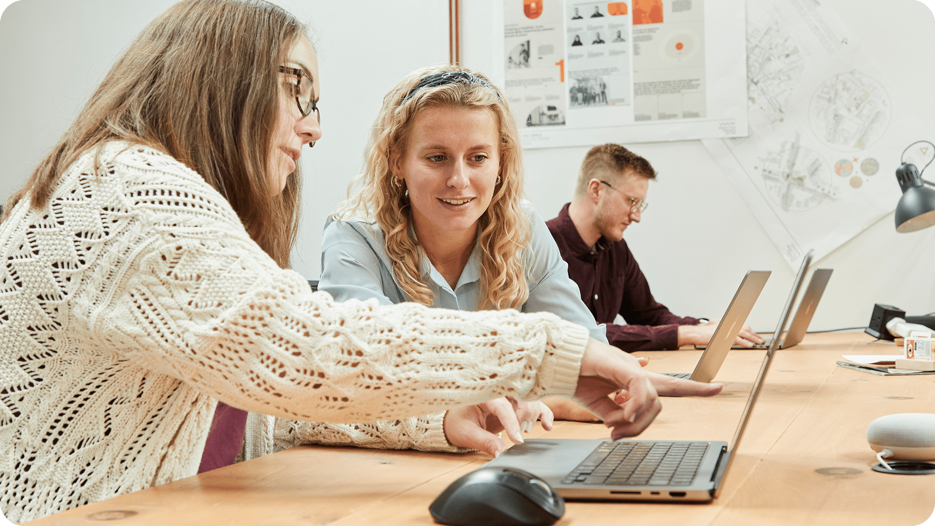 Two women sit at a desk, collaborating and pointing at a laptop screen, while a man works independently on his laptop in the background. The setting appears to be a modern, casual office or classroom.