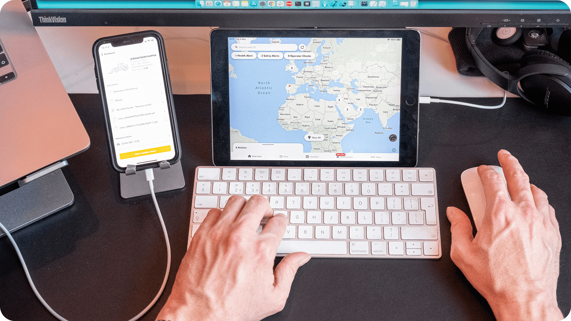 A person uses a wireless keyboard and mouse with a tablet displaying a world map, while a smartphone on a stand shows a ride-sharing app. A laptop and headphones are also visible on the desk.