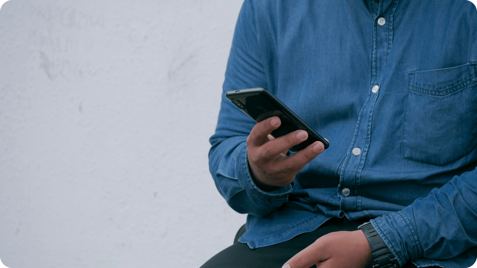A person in a blue denim shirt sits and looks at a smartphone held in one hand, with their other hand resting on their leg. The background is a plain, light-colored wall.