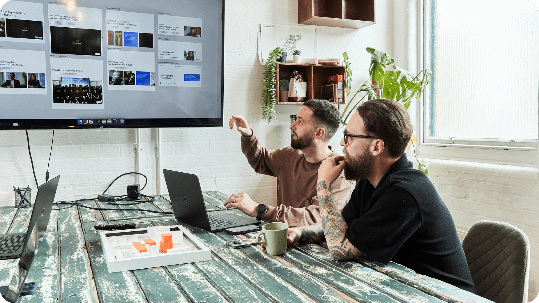 Two people sit at a rustic wooden table with laptops, looking at a large screen displaying a website. One gestures toward the screen while the other watches attentively. Plants and shelves decorate the bright office space.