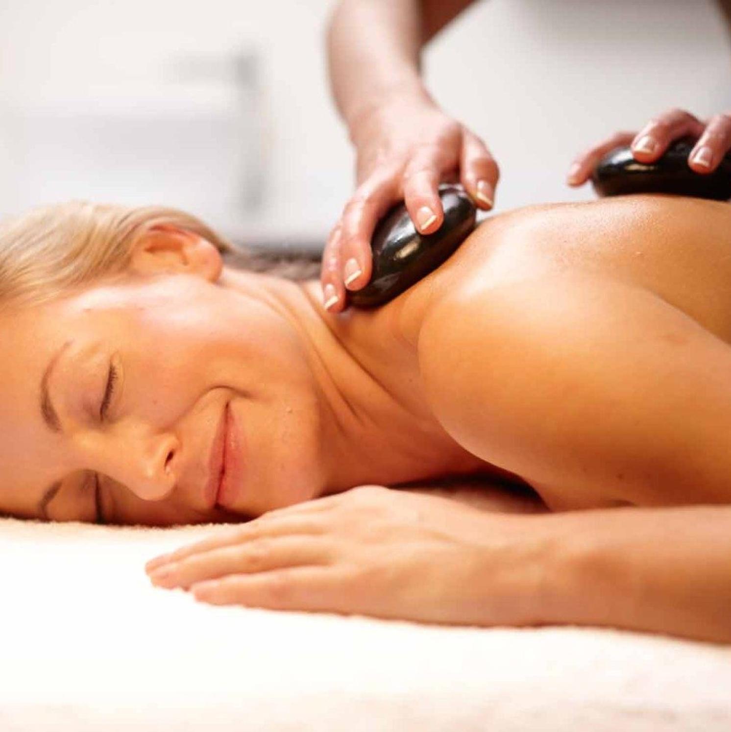 A woman lies face down on a massage table, smiling with her eyes closed, while a person places smooth hot stones on her upper back during a relaxing spa treatment.