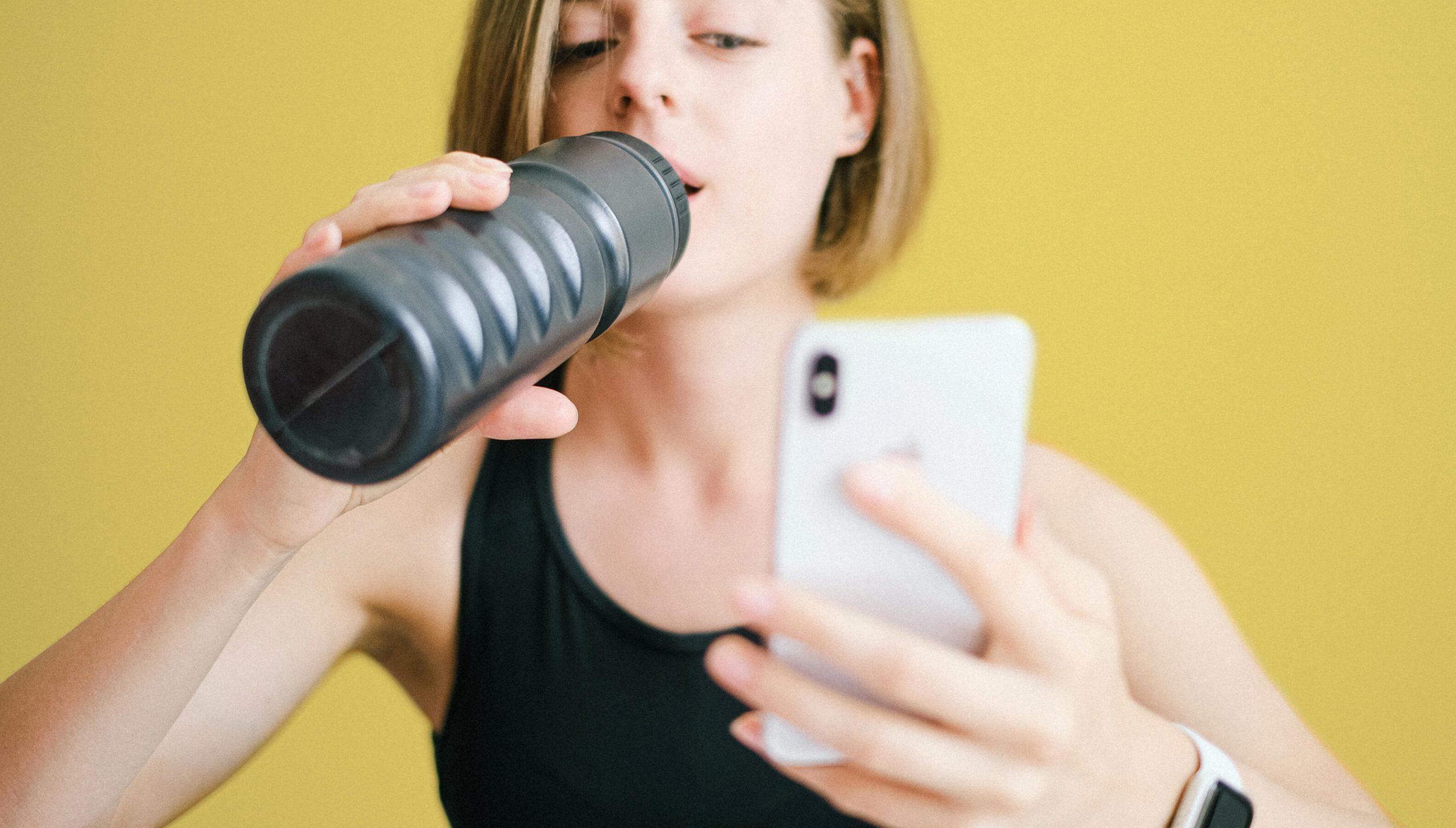 A person in a black tank top drinks from a black water bottle while holding a smartphone, standing against a bright yellow background.
