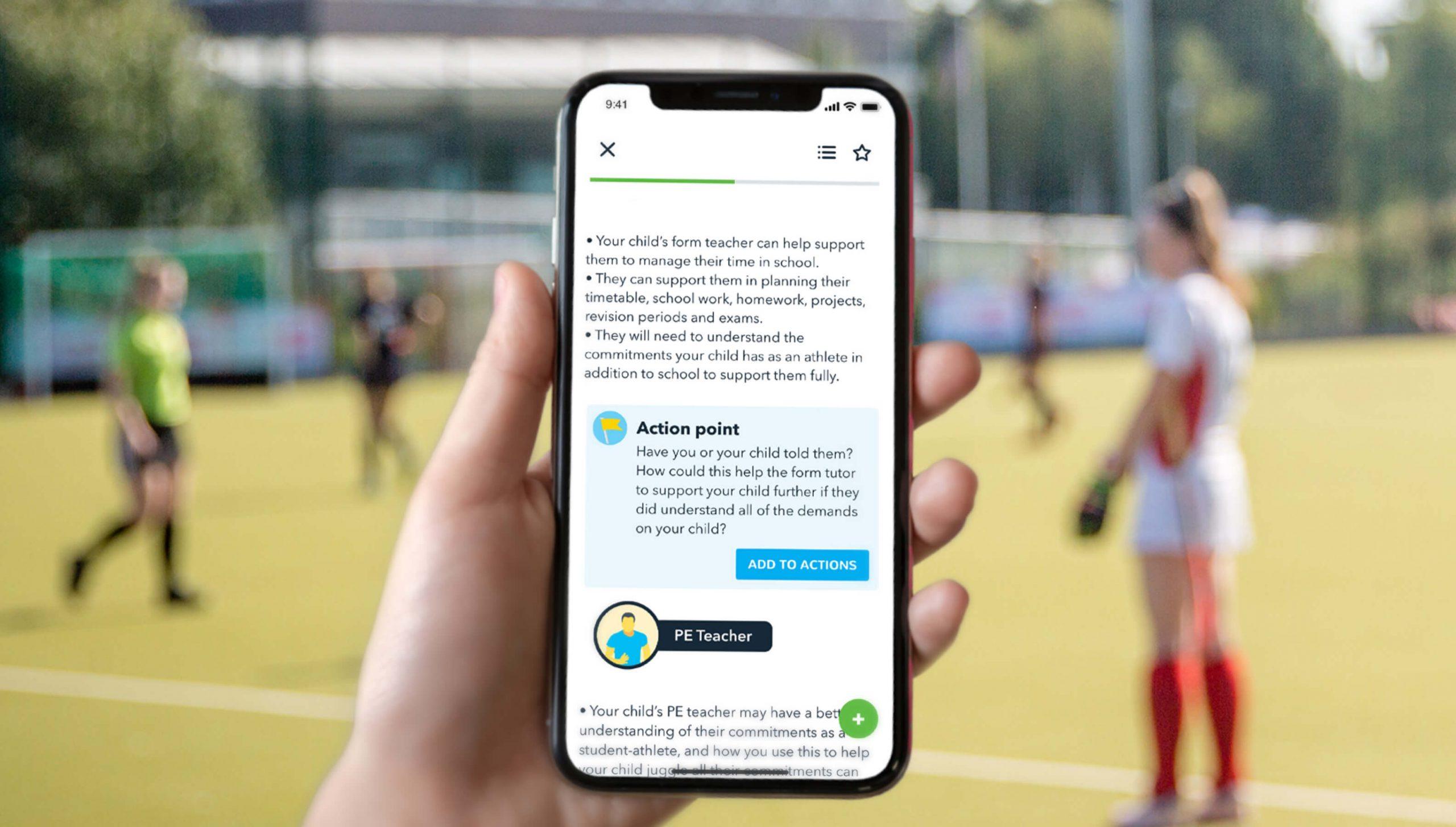 A person holds a smartphone displaying an educational app with a message and an action point from a PE teacher, while a blurred field hockey game takes place in the background.