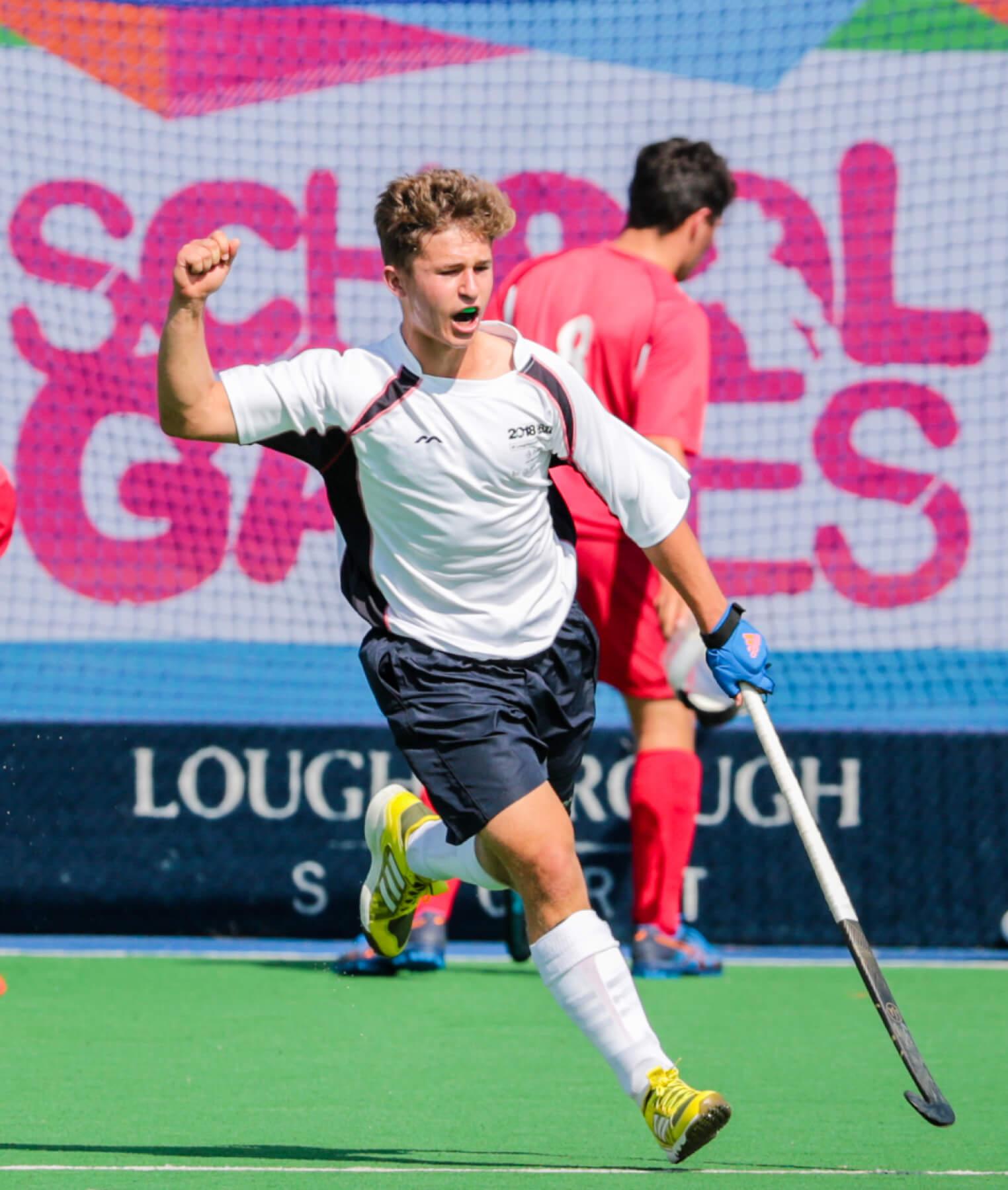 A field hockey player in a white and black uniform celebrates with a raised fist on a green pitch, while a player in a red uniform walks behind. The background shows a colorful banner with School Games text.