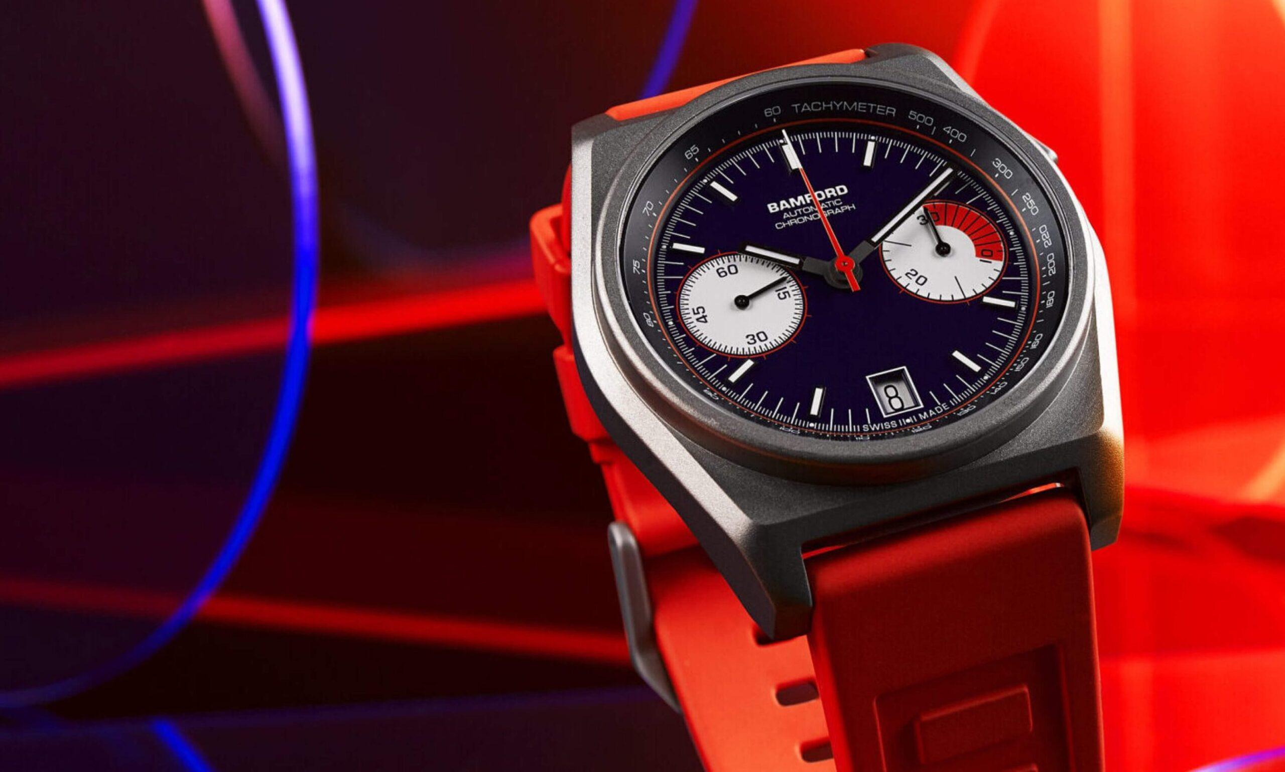 A close-up of a Bamford chronograph watch with a black face, red accents, red strap, and silver bezel, set against a vibrant red and blue abstract background.