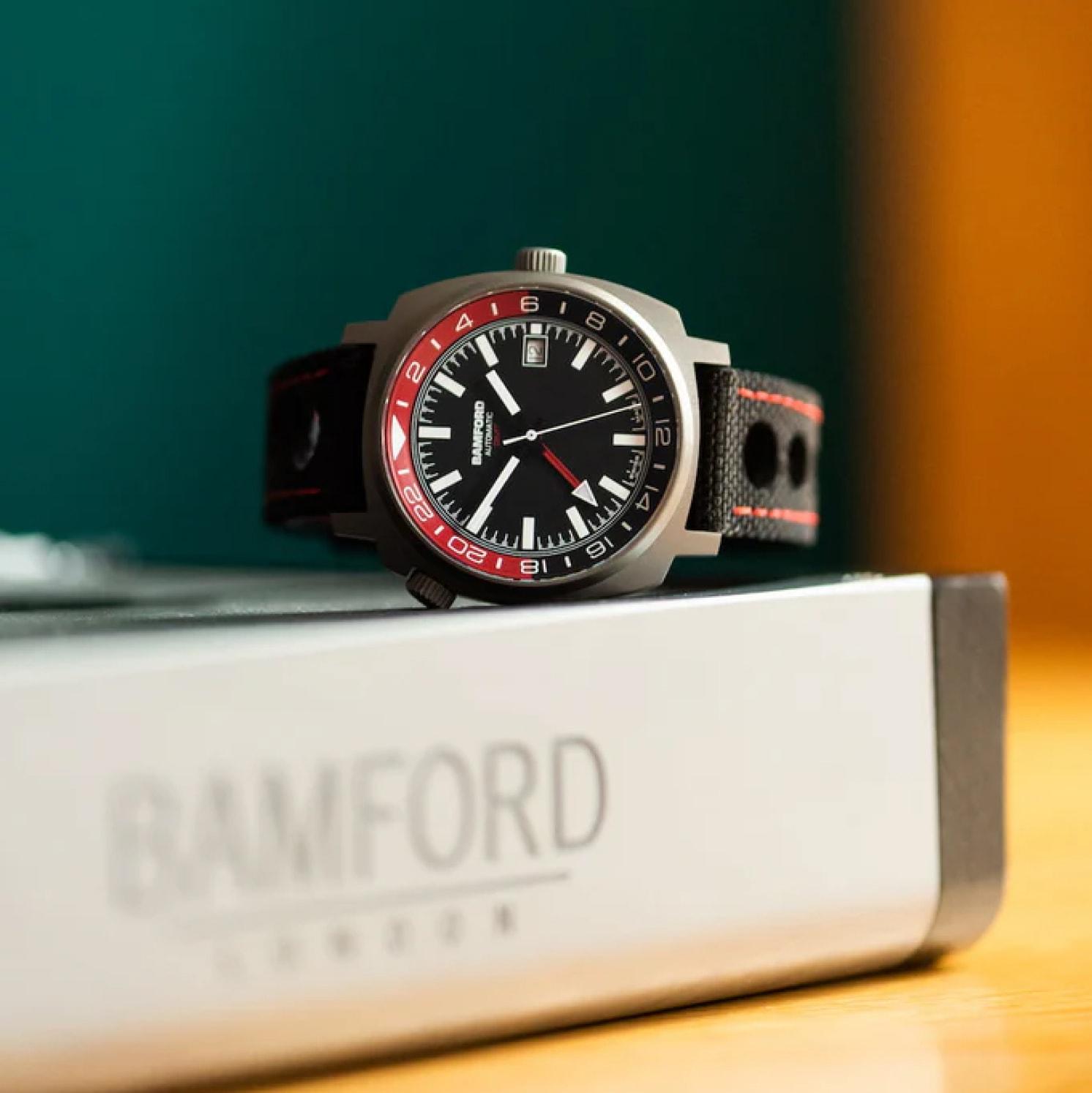A wristwatch with a black dial, red and white accents, and a black leather strap rests on a box labeled BAMFORD London. The background is blurred with green and orange tones.
