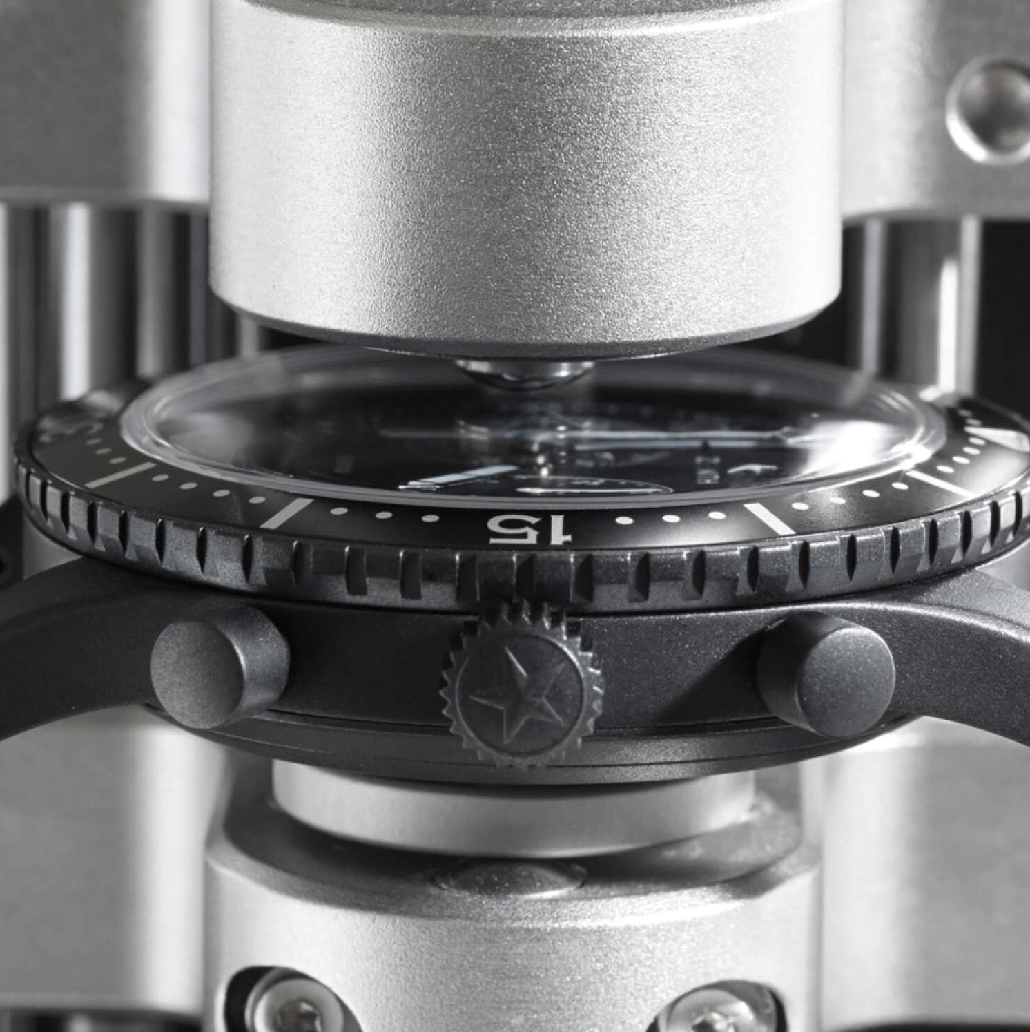 Close-up of a wristwatch case being pressed by a metallic machine, possibly during the assembly or testing process. The watch bezel and crown are visible, with the number 15 showing on the bezel.