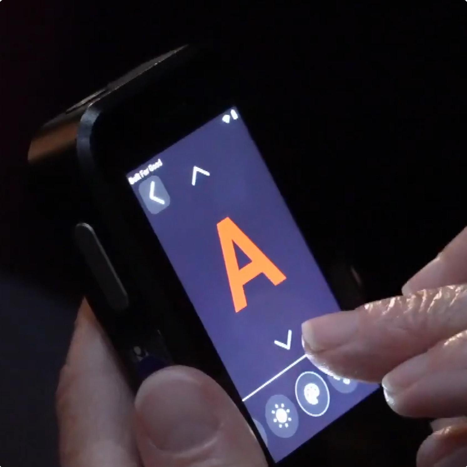 A person uses a touchscreen device displaying a large orange letter A on a dark background, with navigation and brightness icons visible at the bottom of the screen.