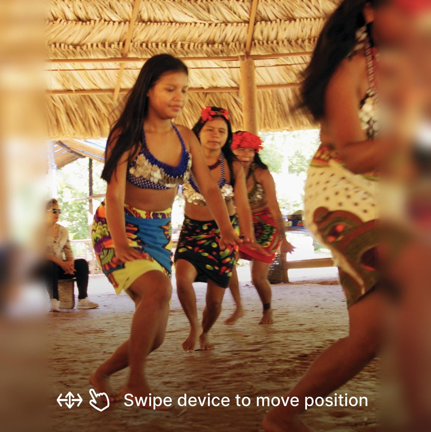 Four women dressed in colorful traditional attire perform a dance in a thatched-roof open structure, with blurred background figures watching. The text reads: Swipe device to move position.
