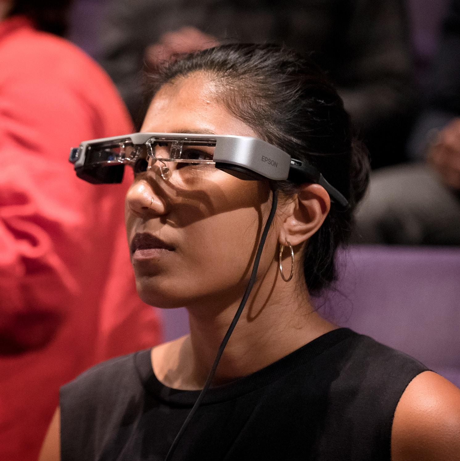 A woman wearing smart glasses with a headset and clear frames, looking ahead in a seated indoor setting with blurred people in the background.