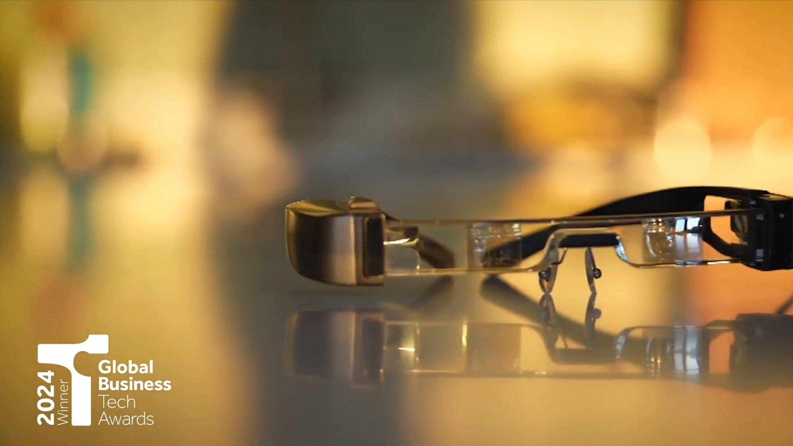 A close-up of smart glasses resting on a reflective surface, with a blurred background. The 2024 Winner Global Business Tech Awards logo is visible in the bottom left corner.