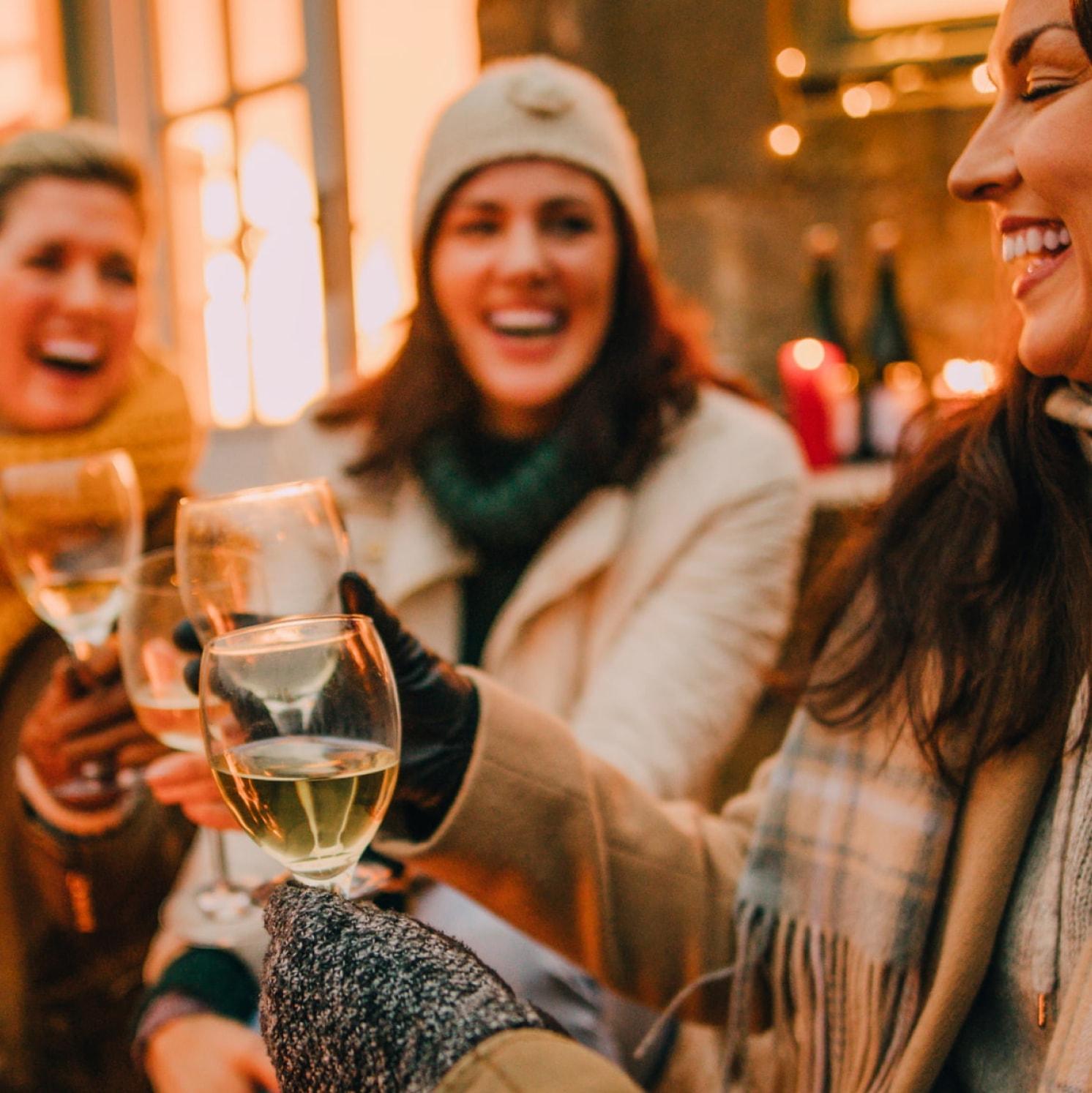 Three women dressed in winter clothing smile and raise glasses of white wine together outdoors, enjoying a cheerful moment in warm, cozy lighting.