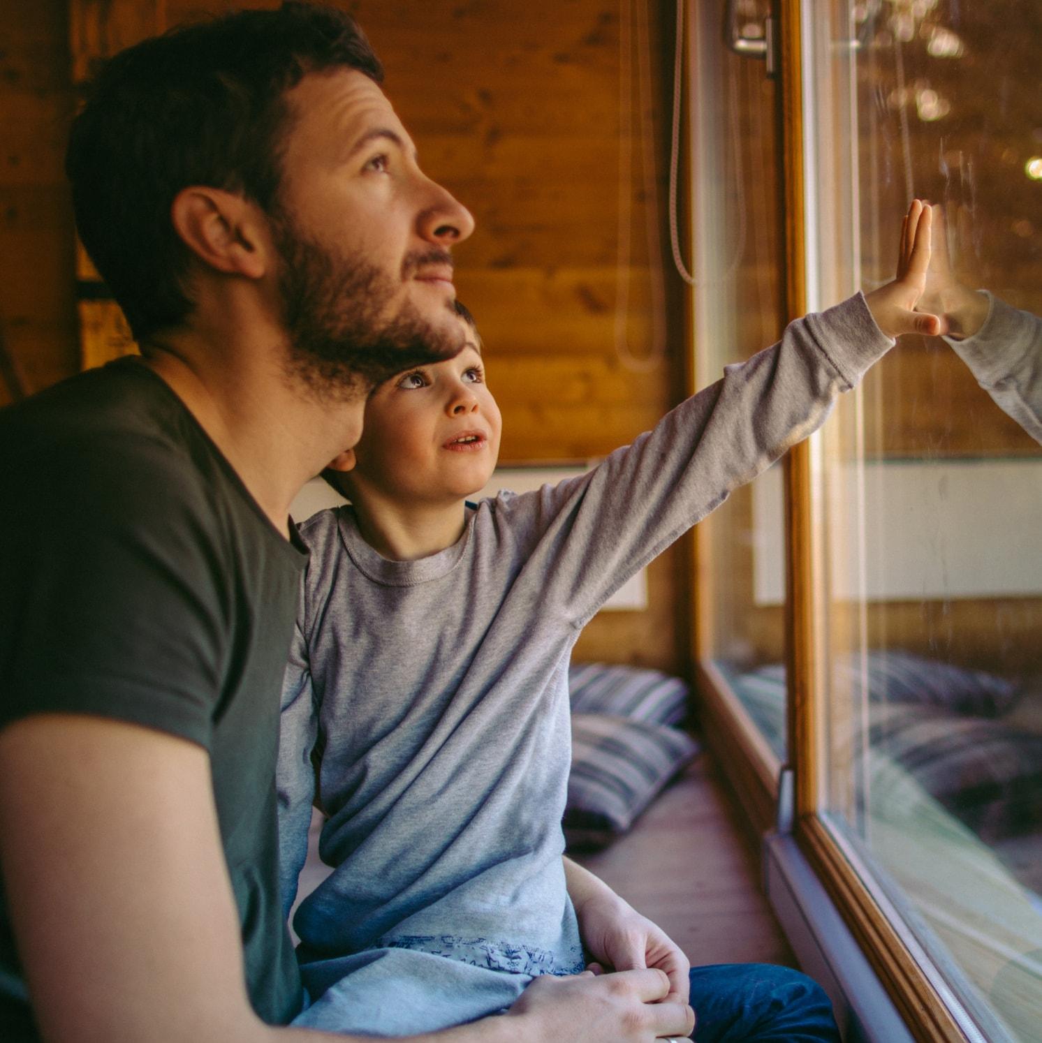 A man and a young boy sit by a large window in a cozy wooden room. The boy touches the glass and gazes outside with curiosity, while the man looks out thoughtfully, holding the boy on his lap.