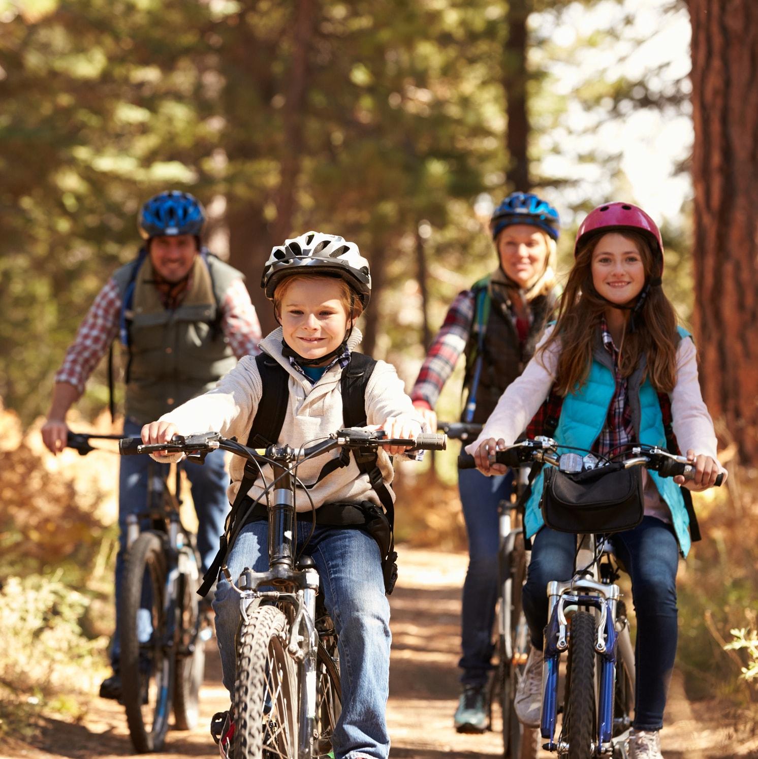 A family of four wearing helmets rides bicycles on a forest trail. Two children are in front, smiling, with two adults behind them. Sunlight filters through the trees, and everyone looks happy and active.