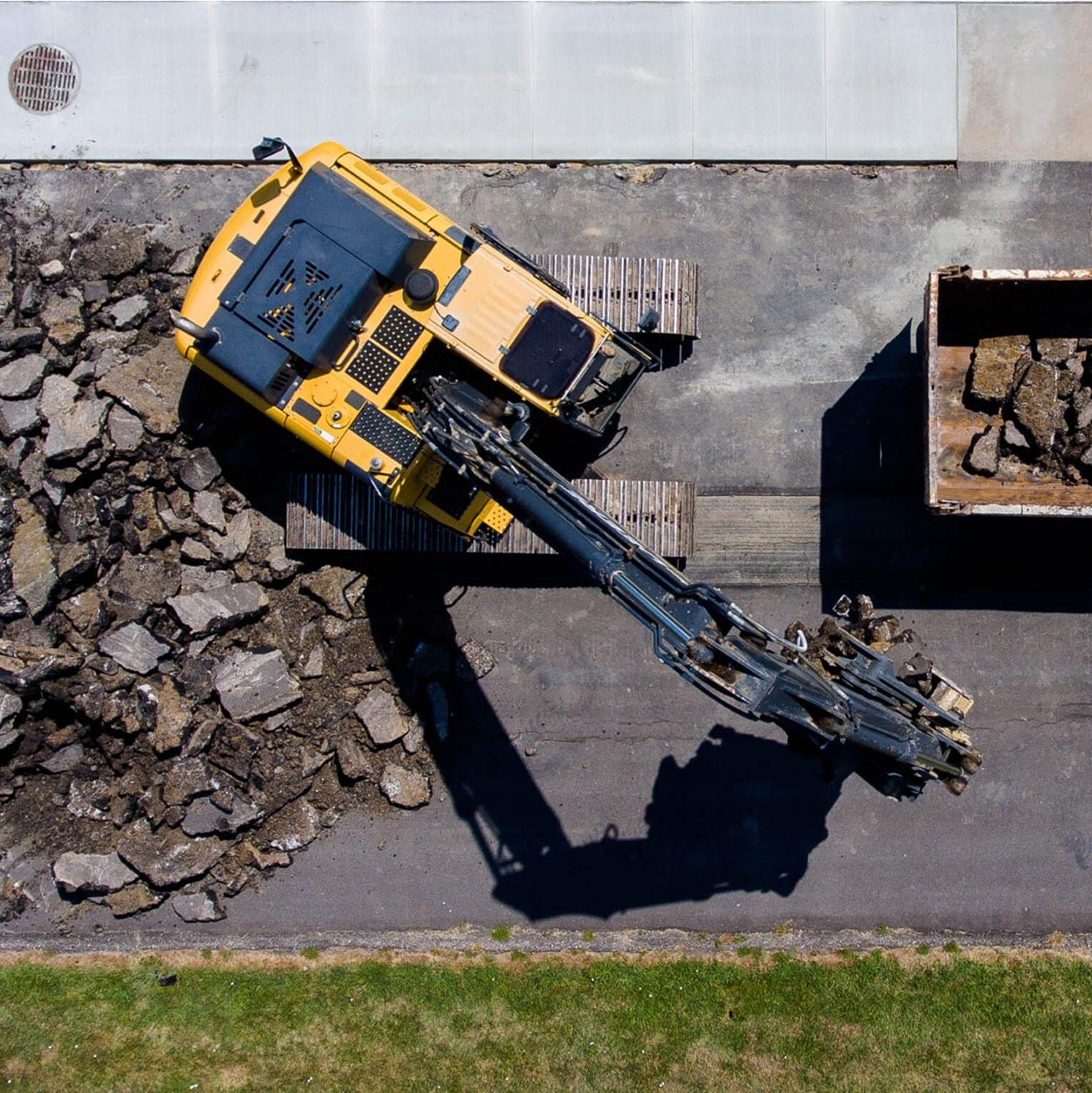 Aerial view of a yellow excavator breaking up asphalt on a paved surface, with large pieces scattered around. The arm of the excavator extends toward a nearby dumpster partially filled with debris.