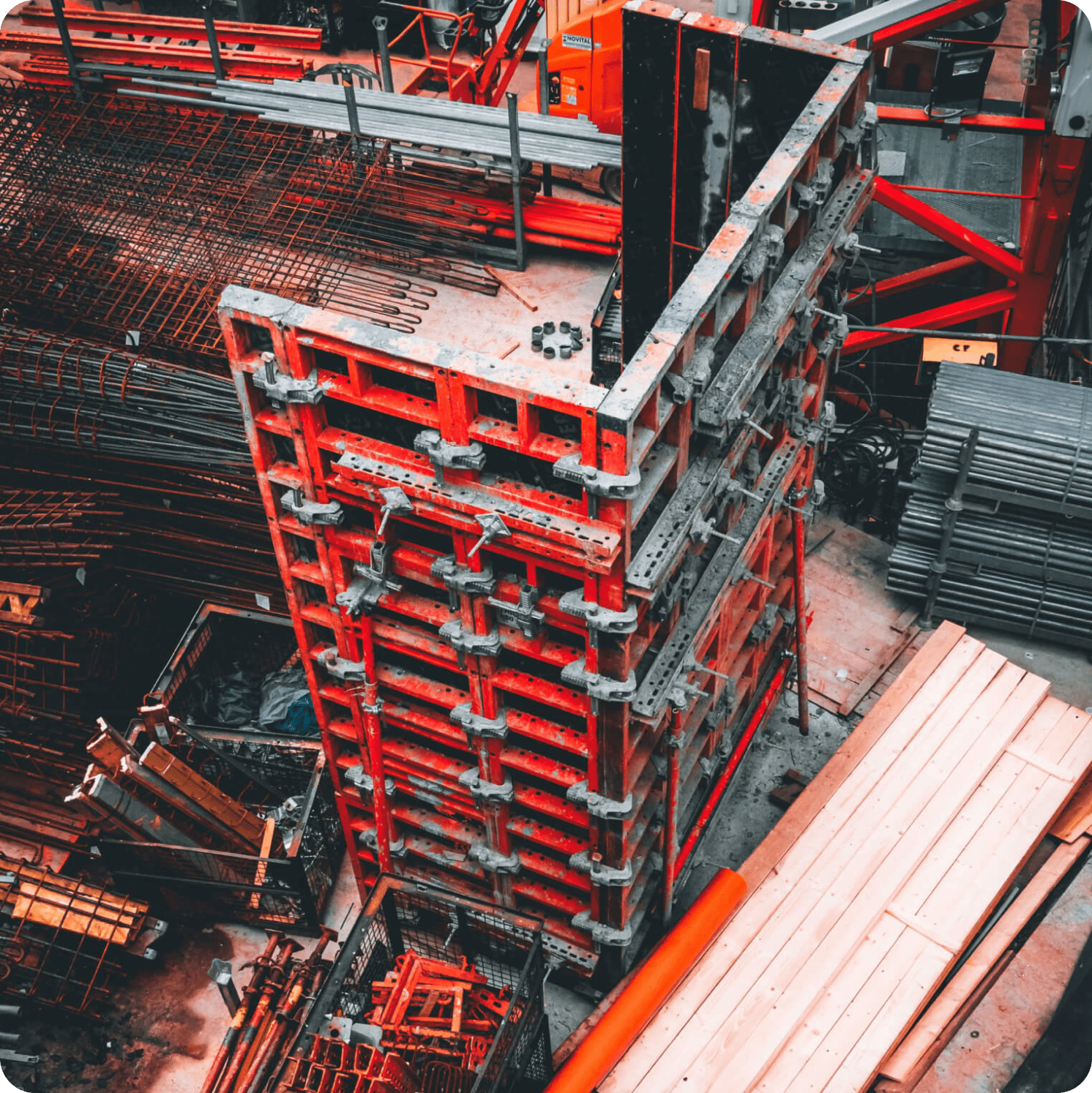 A construction site with stacked red and gray metal formwork, metal rods, wooden planks, and scattered building materials arranged around the site.