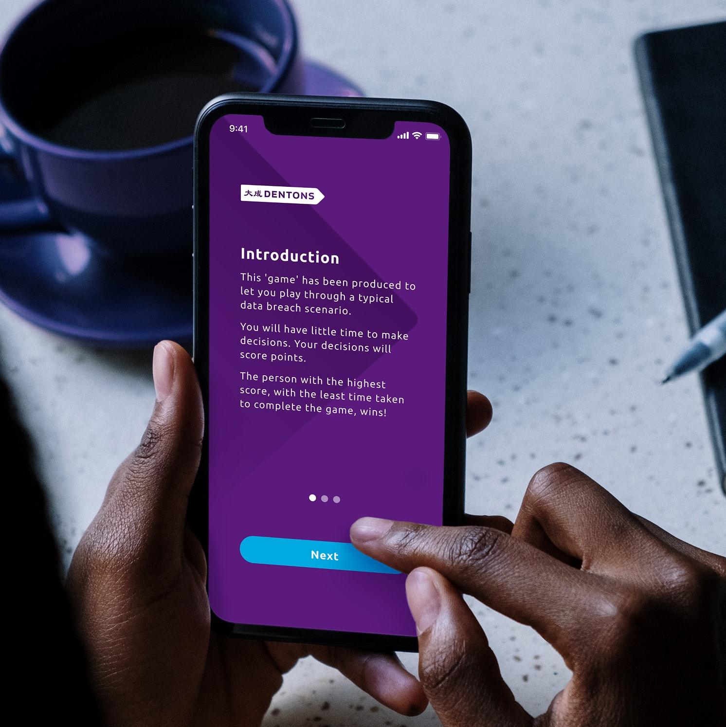 A person holds a smartphone displaying an app with a purple screen titled Introduction, describing a data breach scenario game. Their finger is about to tap a blue Next button. A cup and saucer are in the background.