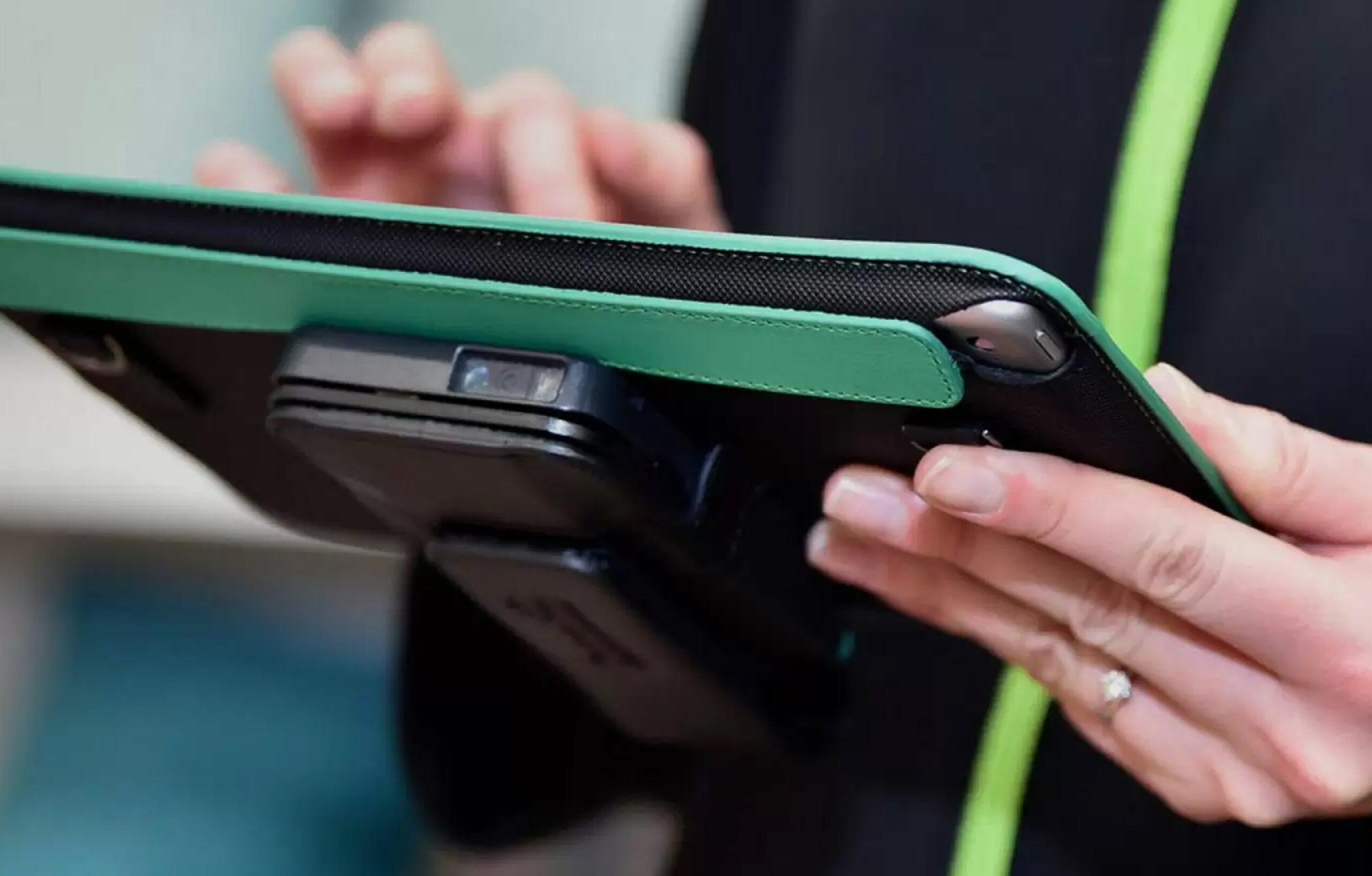 A person holding a tablet with a green case, using their fingers to interact with the screen; a ring is visible on one finger, and the tablet has an attached black device on the back.