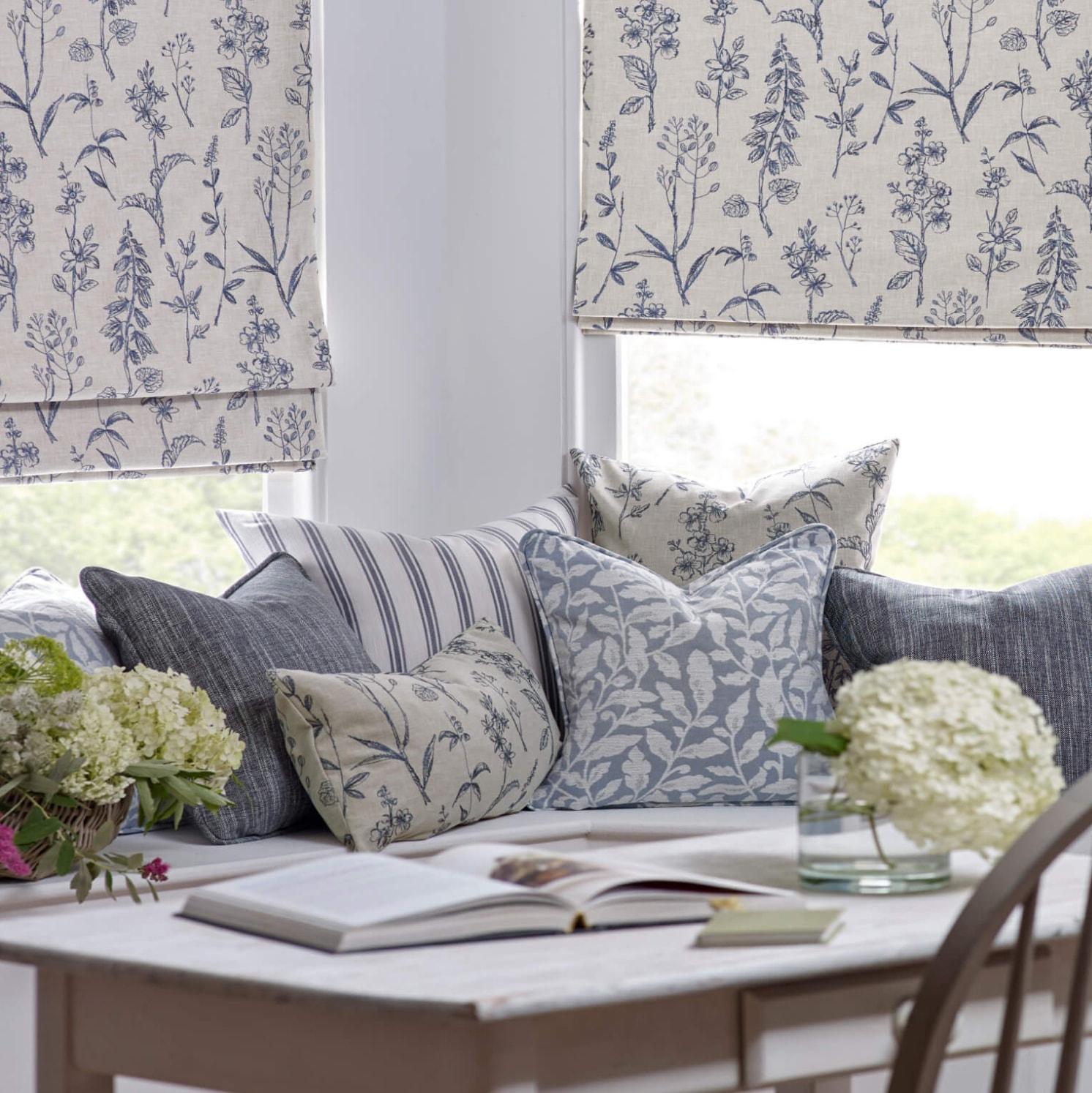 A cozy window seat with patterned cushions and Roman blinds, all featuring blue and white botanical designs. A table in front holds a vase of white hydrangeas and another of green flowers. Natural light fills the space.