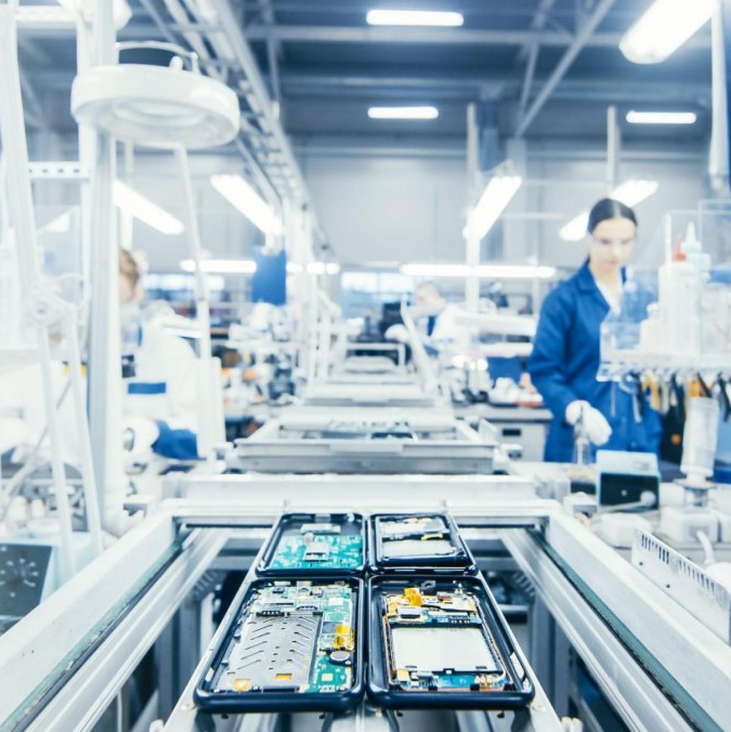 Open smartphones with exposed circuit boards move along a conveyor belt in a brightly lit electronics manufacturing facility, with workers in blue uniforms visible in the background.