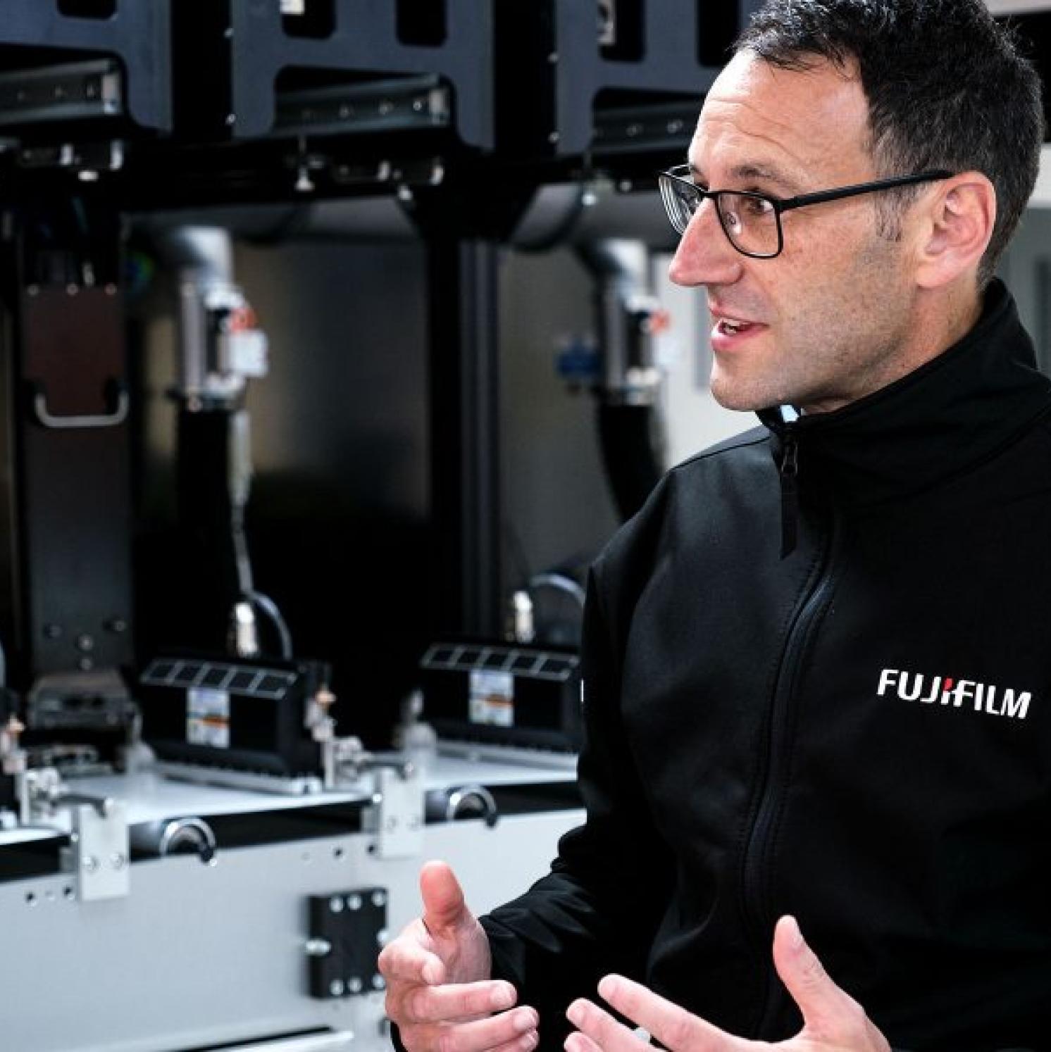 A man wearing glasses and a black FUJIFILM jacket gestures while speaking, standing in front of complex industrial or printing equipment.