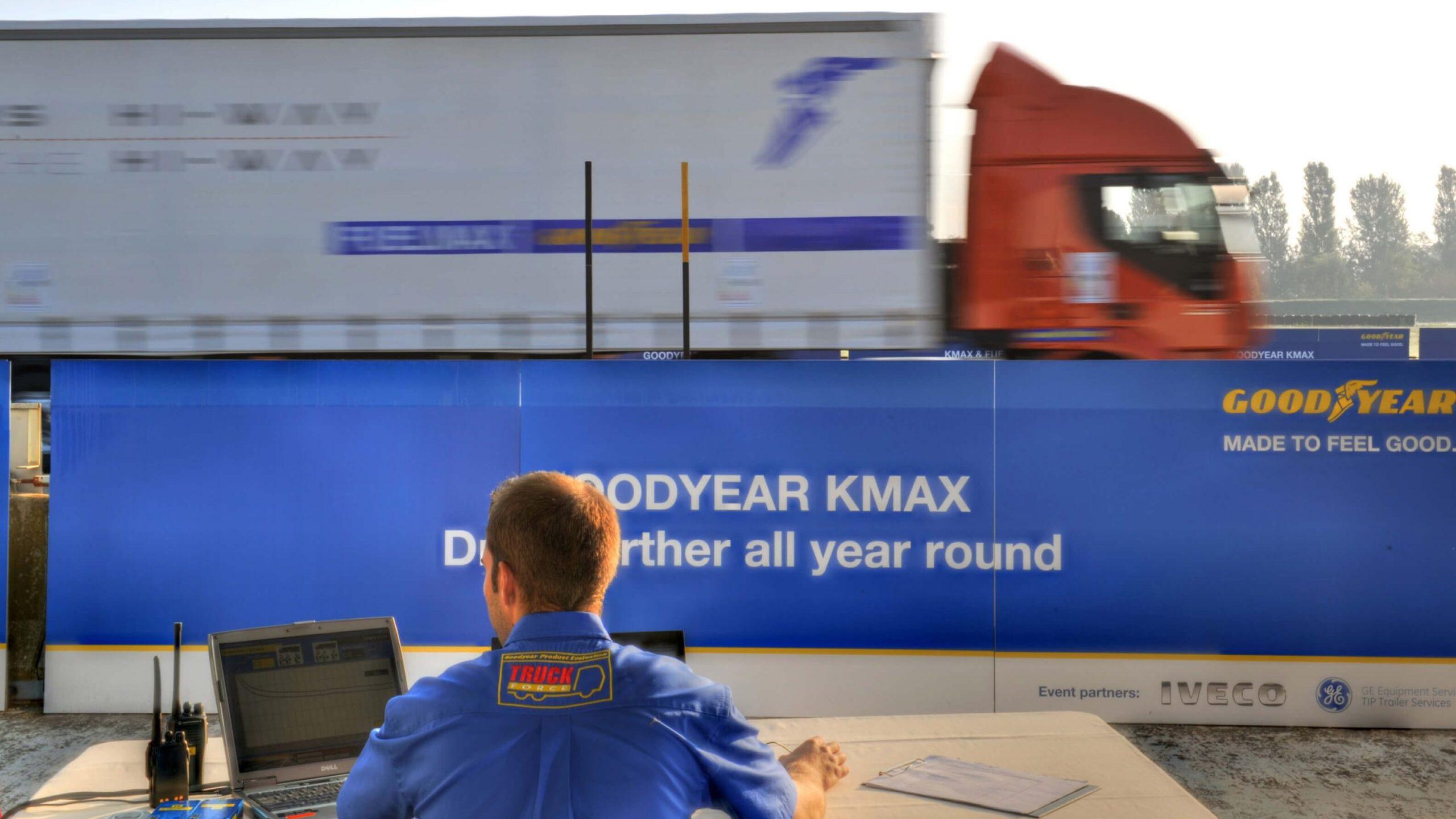 A man in a blue jacket works at a table with a laptop and papers in front of a Goodyear KMAX advertisement, as a red truck with a trailer speeds past in the background.