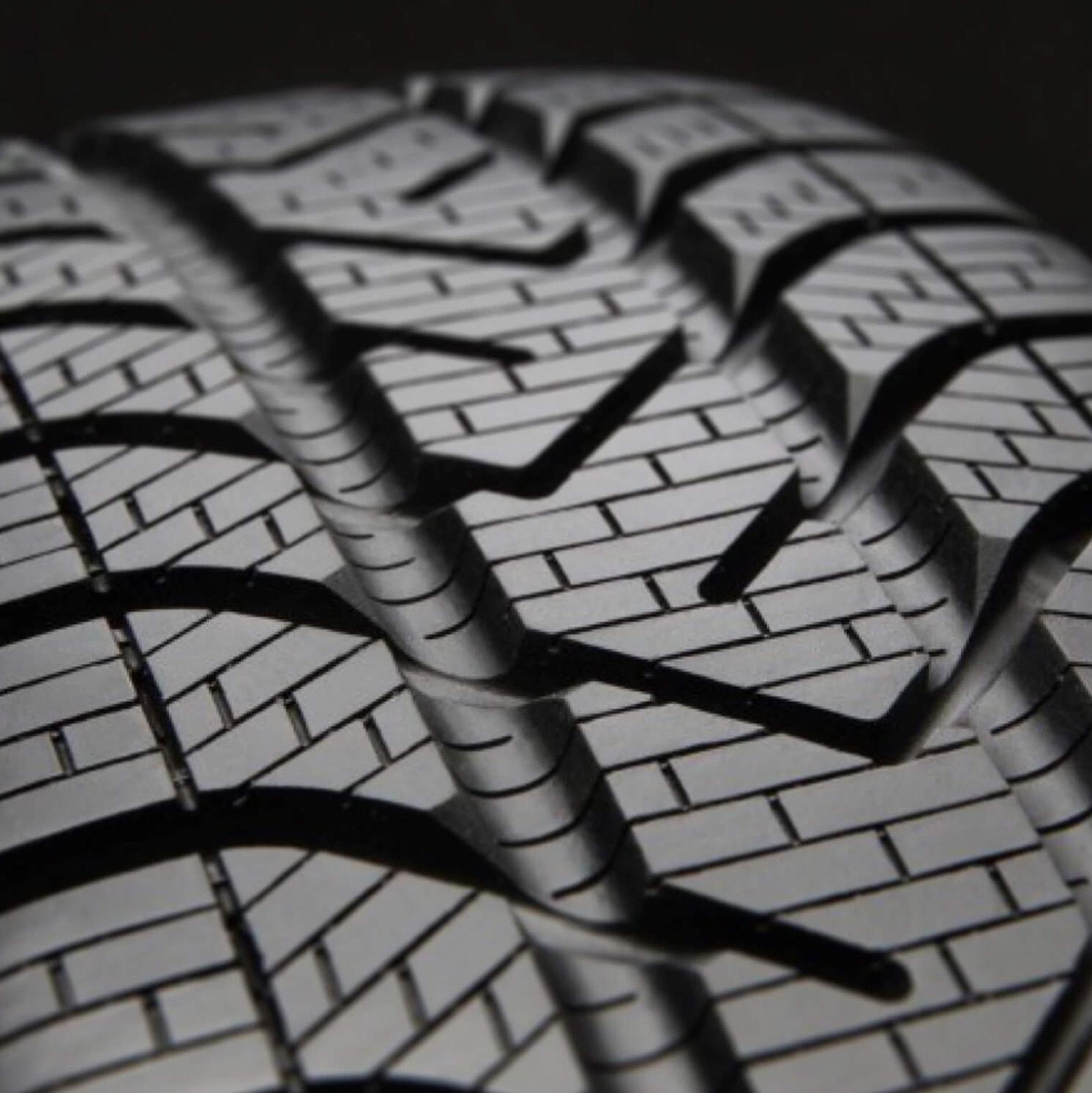 Close-up of a car tire tread pattern, showing the detailed grooves and channels designed for traction and grip on the road. The image highlights the texture and design of the tire surface.