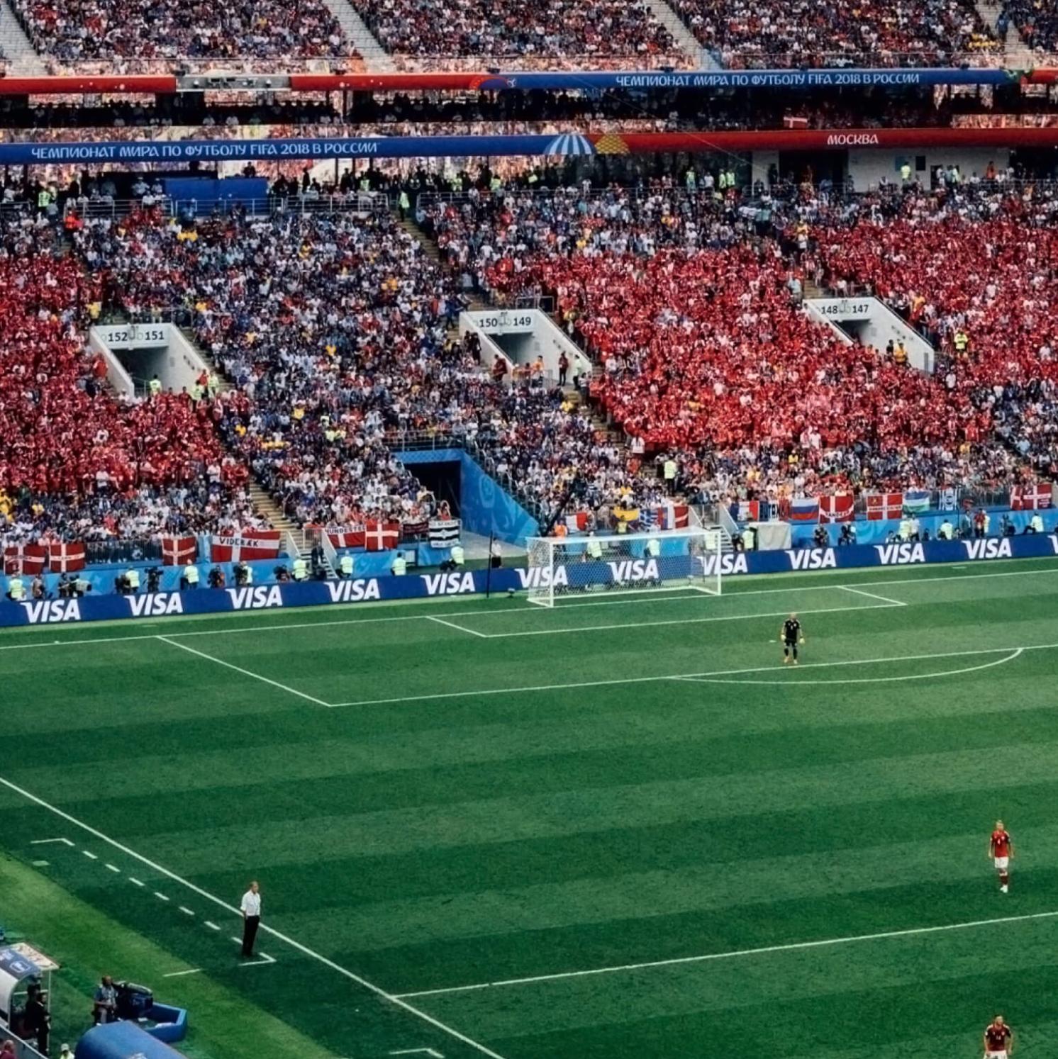 A crowded soccer stadium with fans filling the stands, many holding red and white flags. Two teams are on the green field, with Visa advertisements lining the edges. A few security staff stand near the sidelines.
