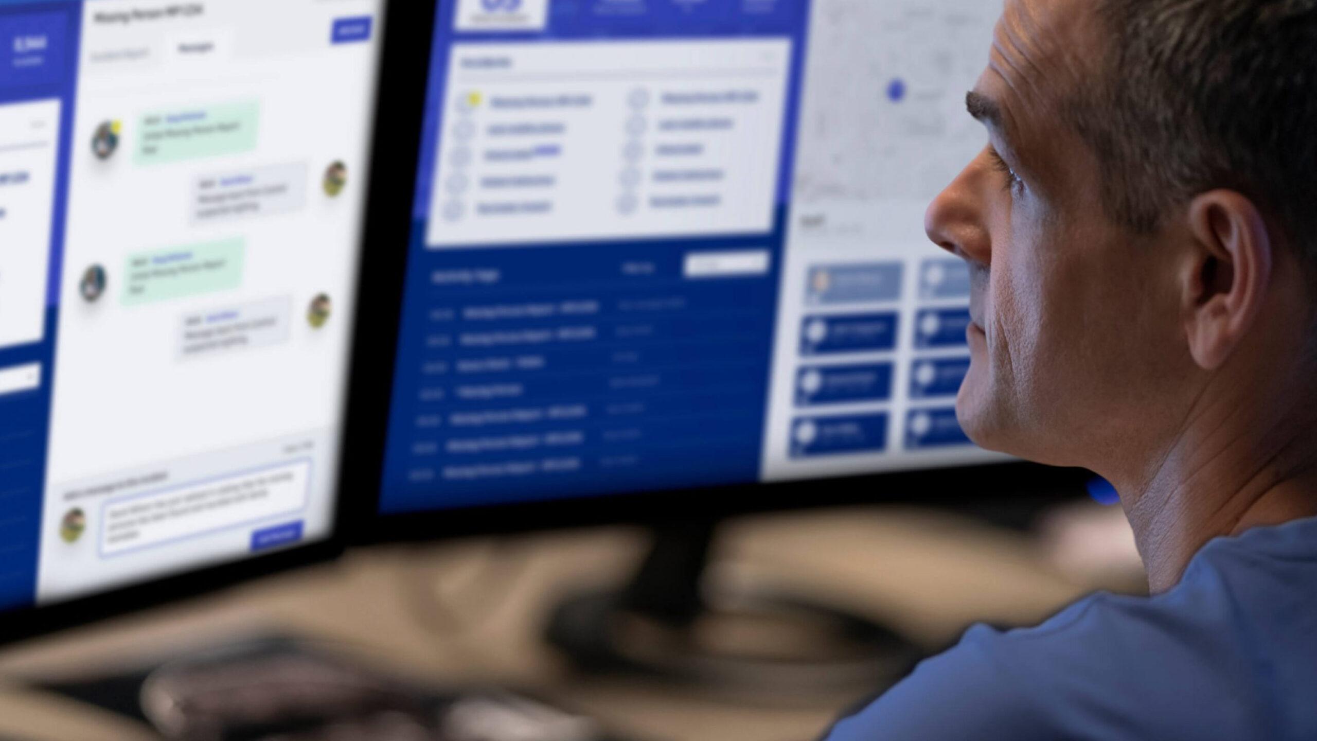 A man looks at two computer monitors displaying messaging apps and dashboards with blue and white interfaces in an office setting.