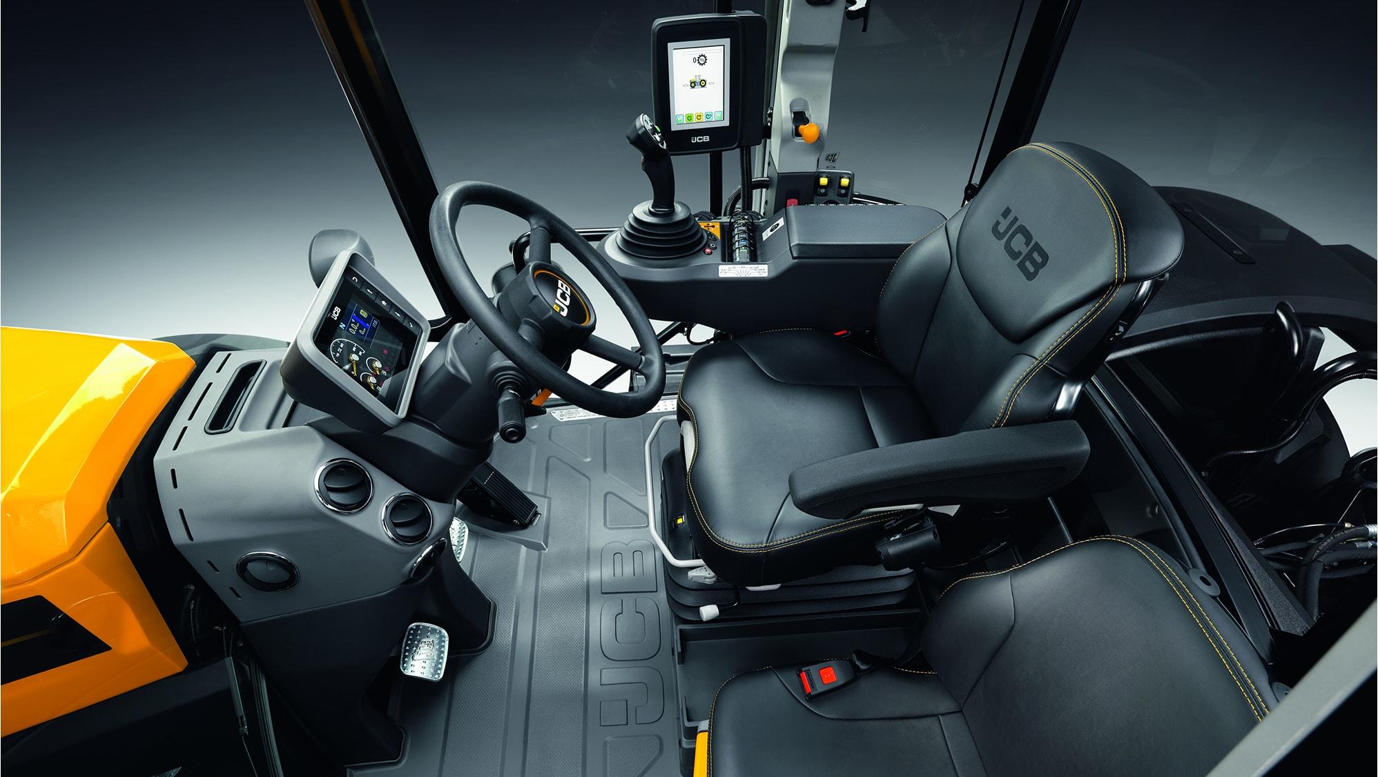 Interior view of a modern JCB construction vehicle cabin, showing the steering wheel, seat, dashboard, control panel with display screen, and various control levers.