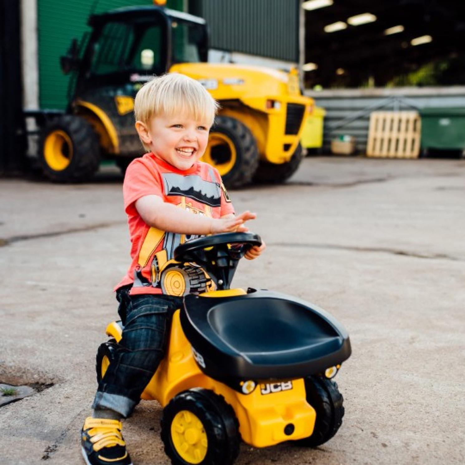 A smiling young boy in a red shirt rides a small yellow toy JCB vehicle outdoors, with a real yellow construction vehicle and green building visible in the background.