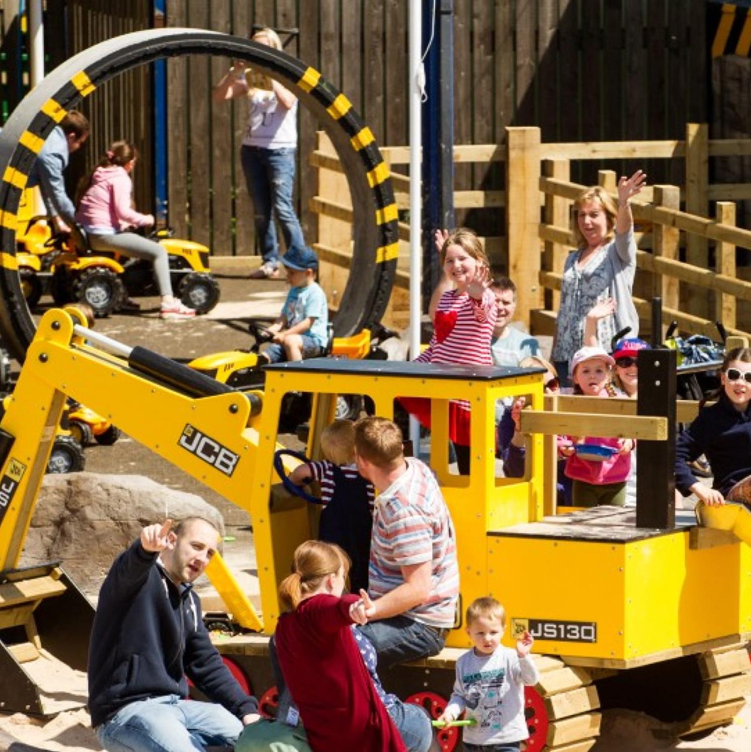 Children and adults play on and around a large yellow digger in an outdoor playground with ride-on toy vehicles and a circular obstacle in the background. Several people are smiling and waving at the camera.