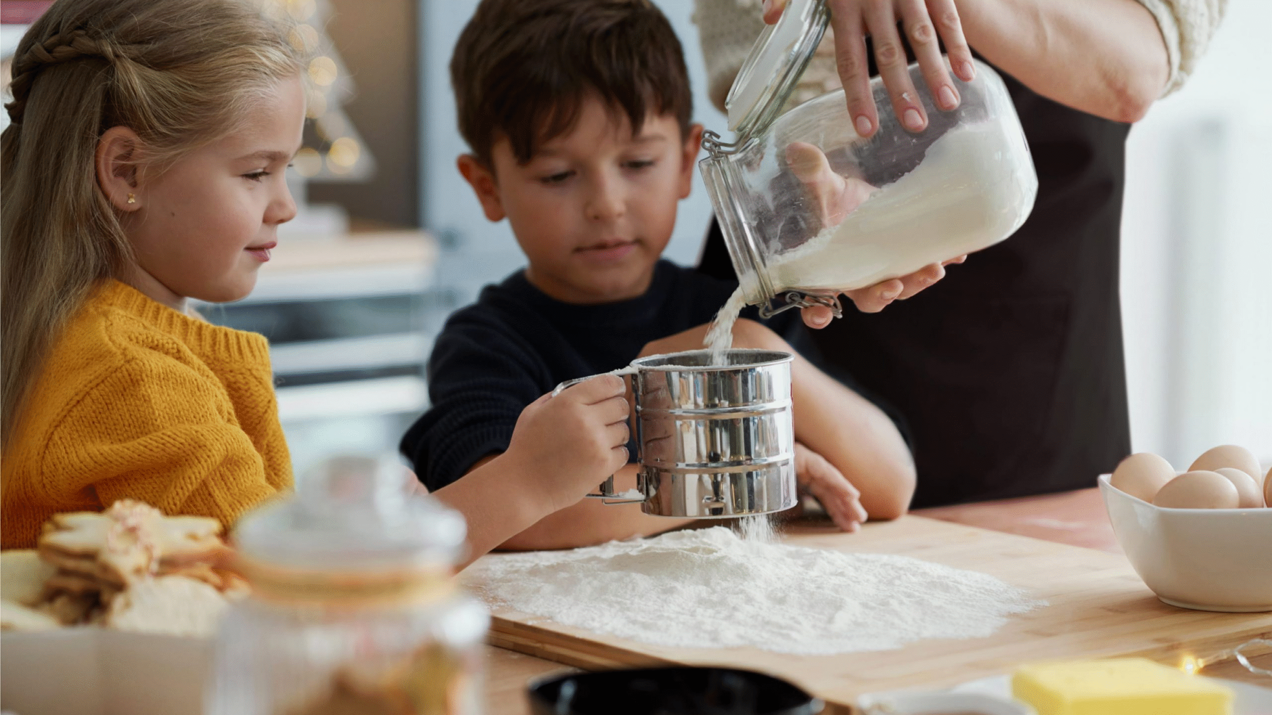 Two children watch as an adult pours flour into a sieve over a floured surface in a kitchen, preparing to bake. Eggs, butter, and biscuits sit nearby on the wooden worktop.