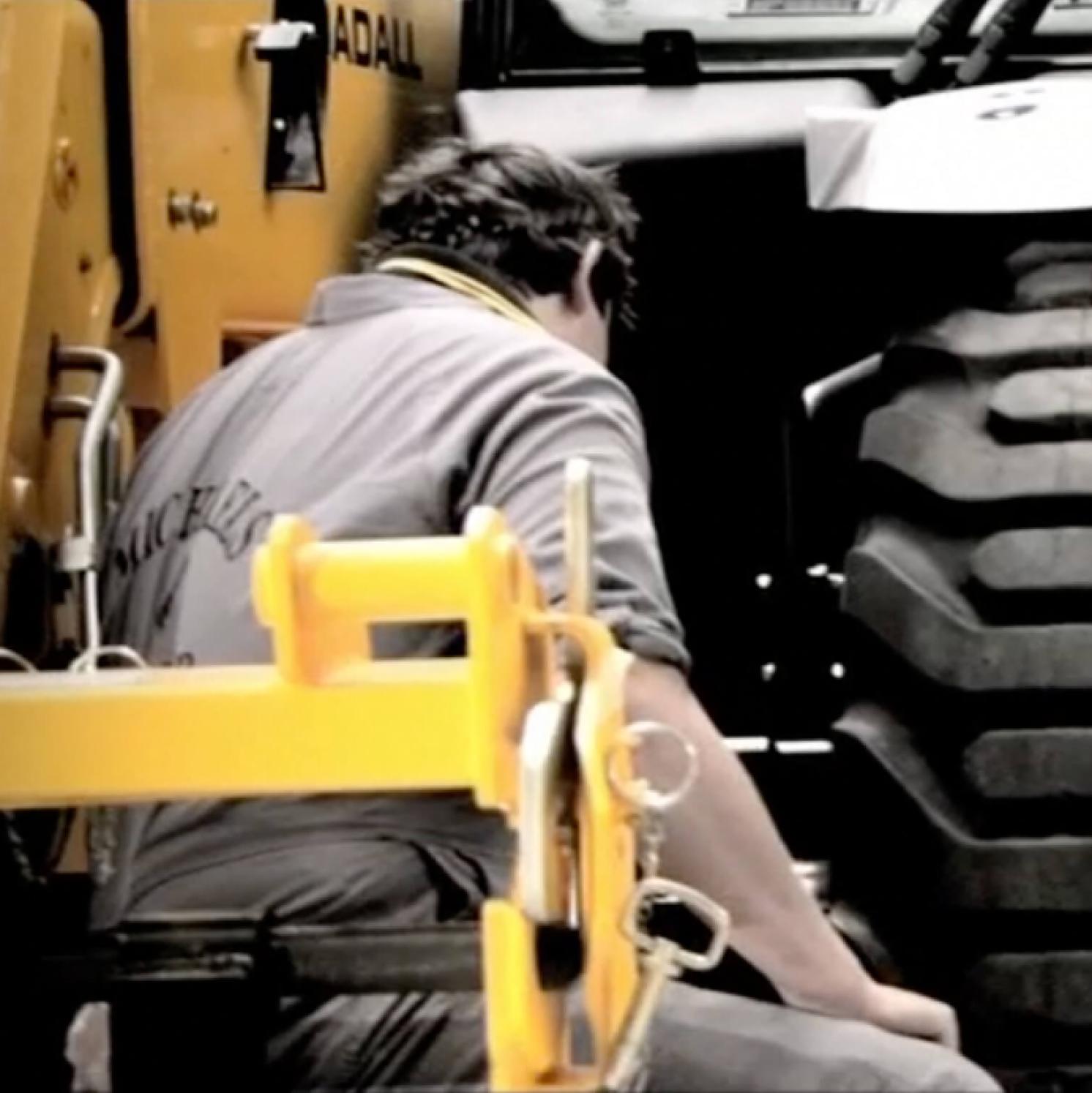 A person in work clothes kneels beside a large yellow construction vehicle, near its black tire, appearing to inspect or repair it. The scene suggests industrial or maintenance work.
