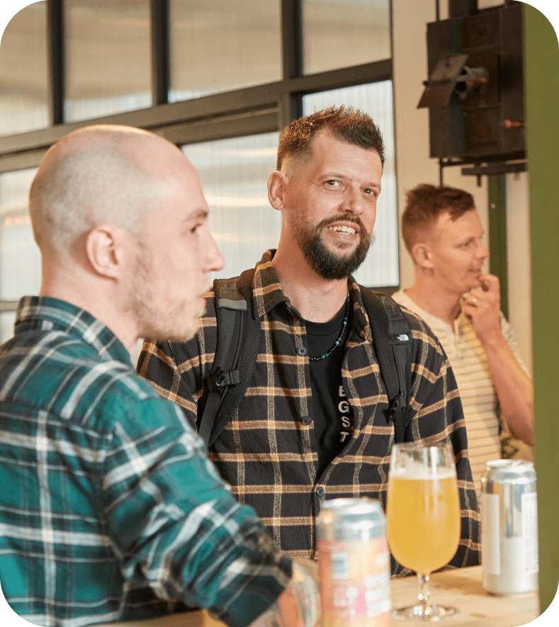 Three men stand and chat at a bar or cafe. Two are in the foreground, both with beards and wearing plaid shirts. The third man in the background wears a striped shirt and holds a glass, while drinks sit on the counter.