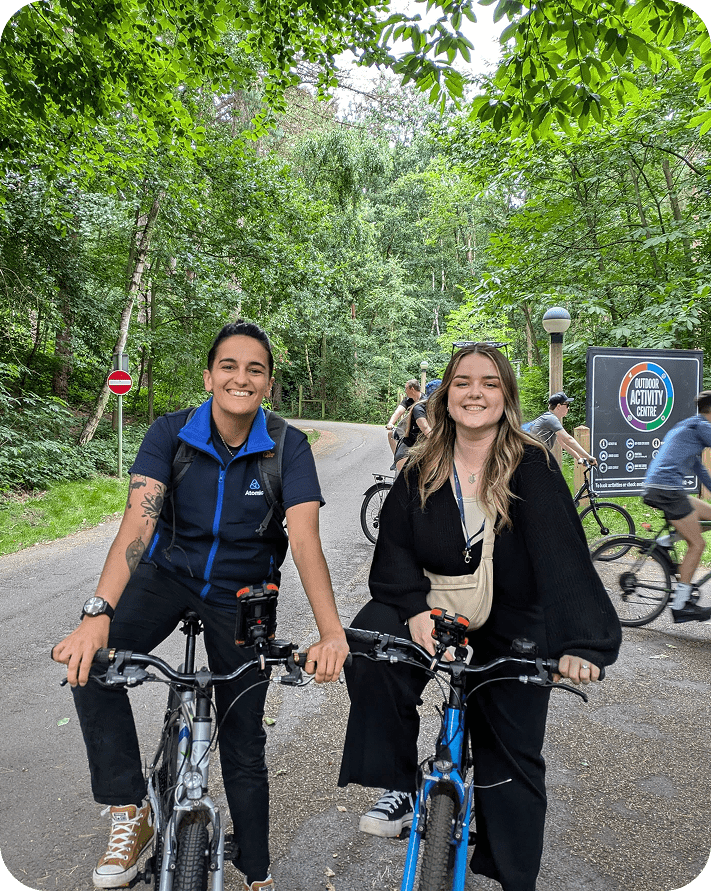 Two people smile while sitting on bicycles on a paved path in a green, wooded park. Other cyclists ride in the background, and a sign with activity information is visible on the right.