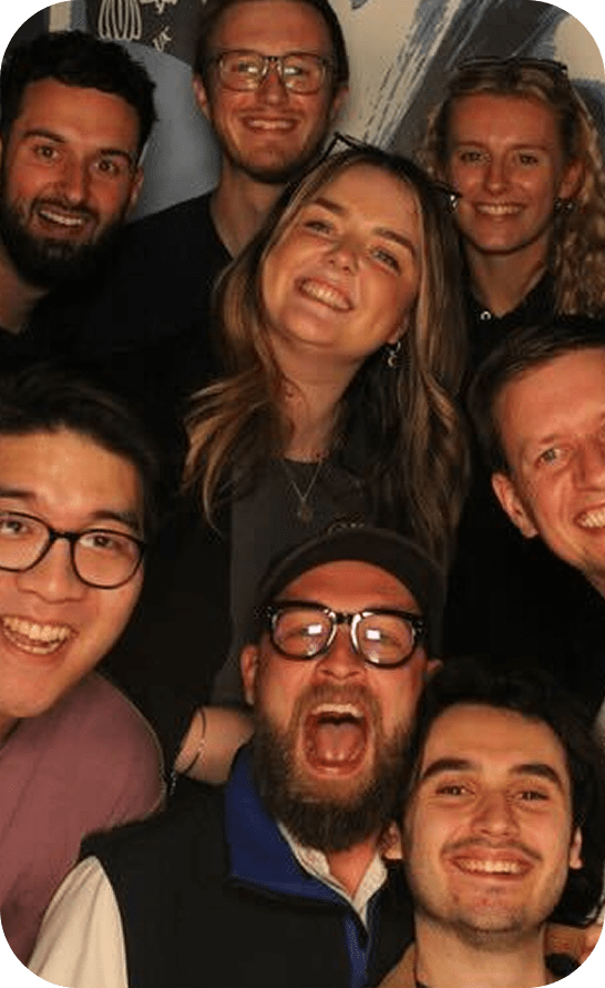 A group of eight people, smiling and laughing, pose closely together for a photo. Most are wearing glasses and casual clothing, and they appear happy and cheerful against a simple background.