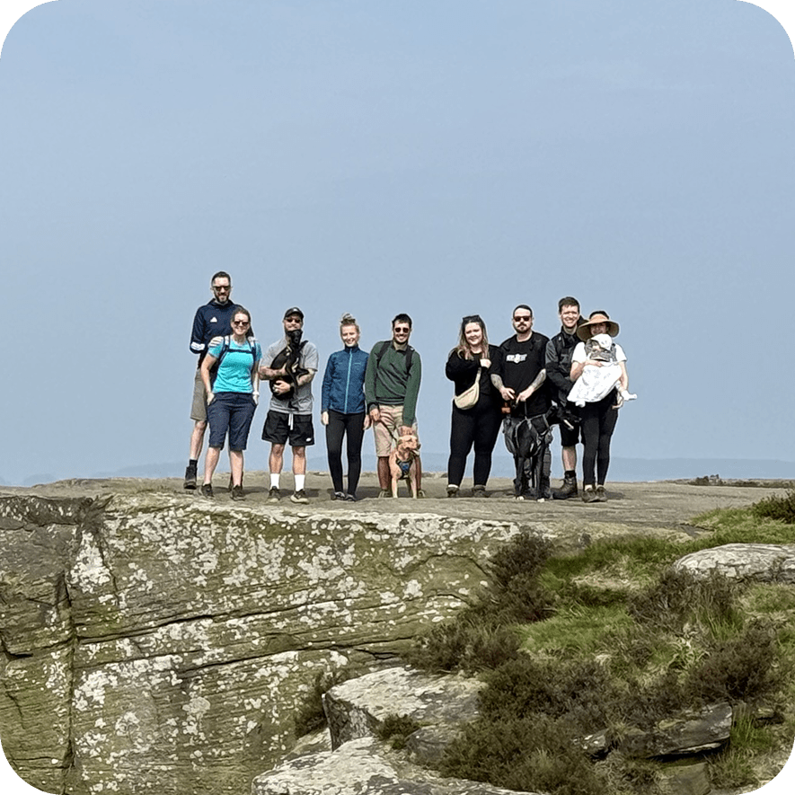 A group of ten people, including a child and a baby, and two dogs stand together on a rocky cliff with grass and shrubs, under a clear blue sky.