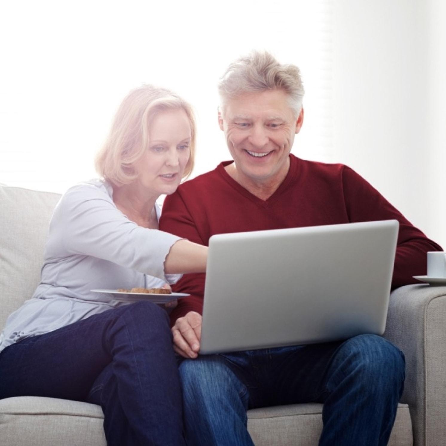 An older couple sits on a couch, smiling and looking at a laptop together. The woman is pointing at the screen while the man navigates, and there is a plate of cookies on her lap. Bright natural light fills the background.