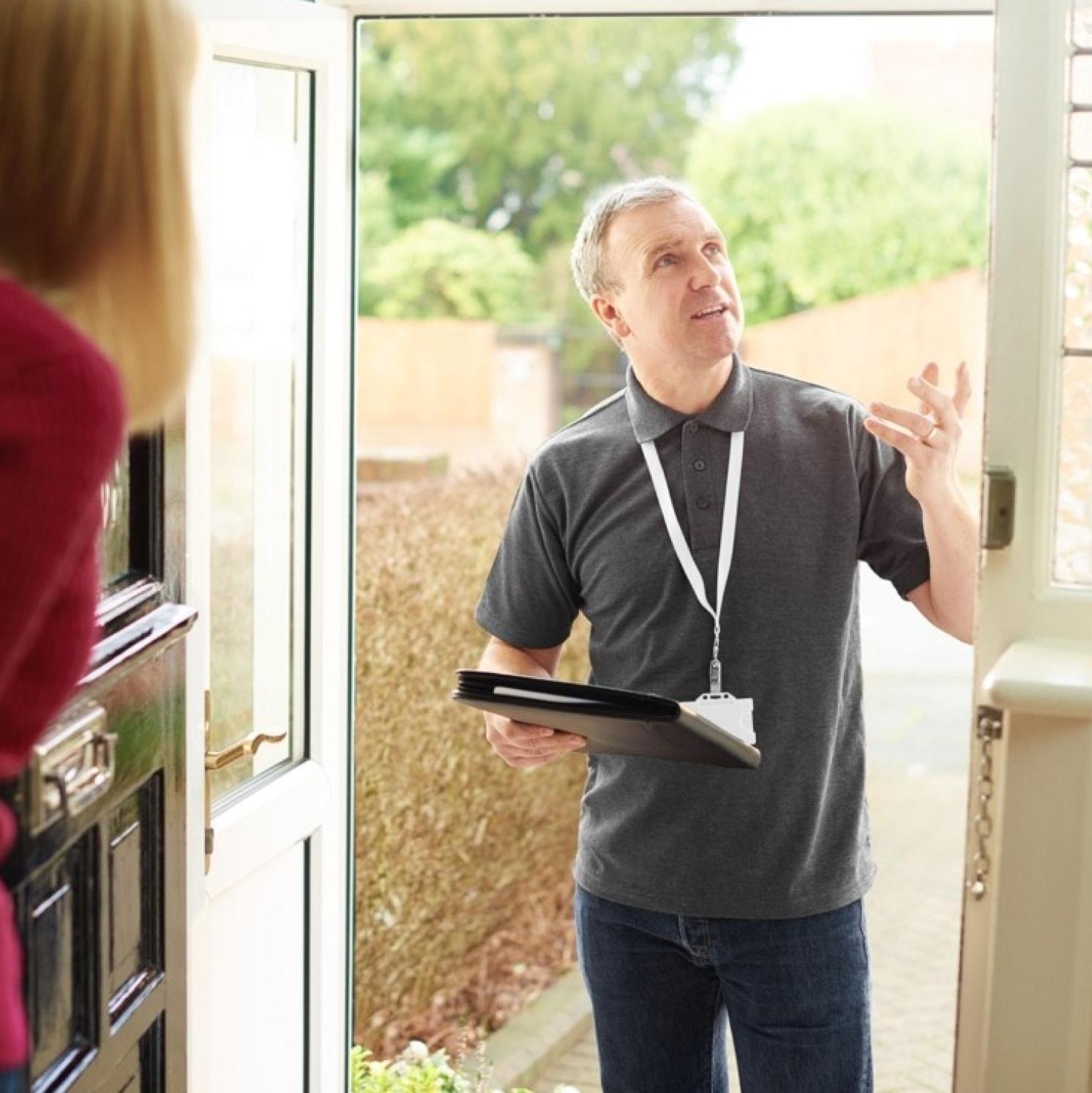 A man with a lanyard and clipboard stands at an open doorway, speaking to a person inside the house. The man gestures upward as he talks, and greenery is visible outside.